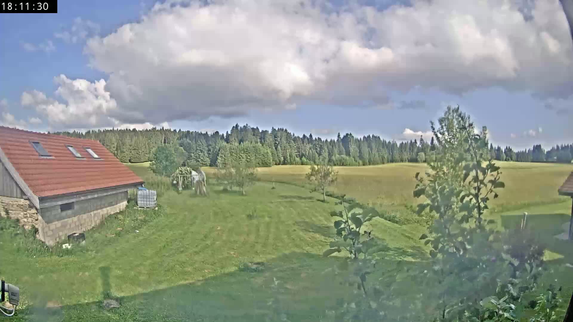 A partly cloudy sky overlooks a grassy yard with a red-roofed building, several trees, and a field beyond a line of trees in the distance.