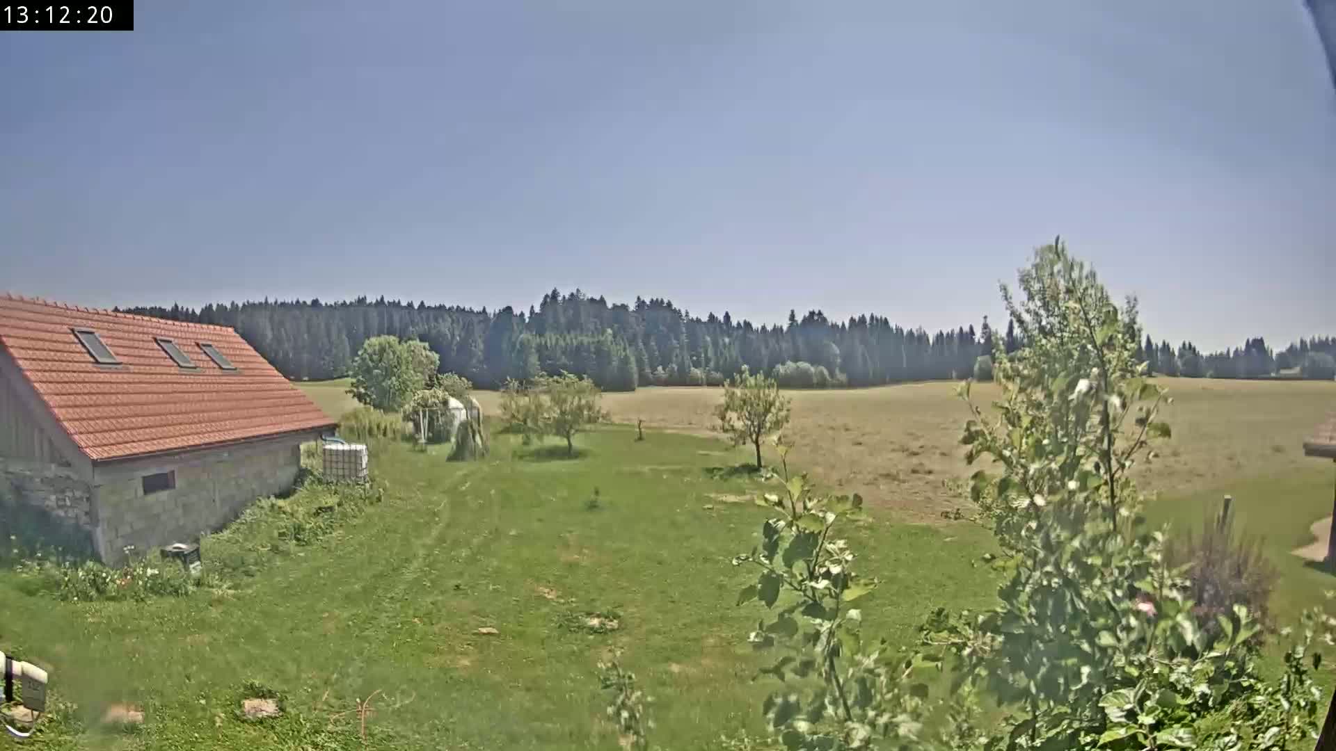A stone building with a red roof sits on a grassy yard, overlooking a dry field and a distant forest under a clear blue sky.