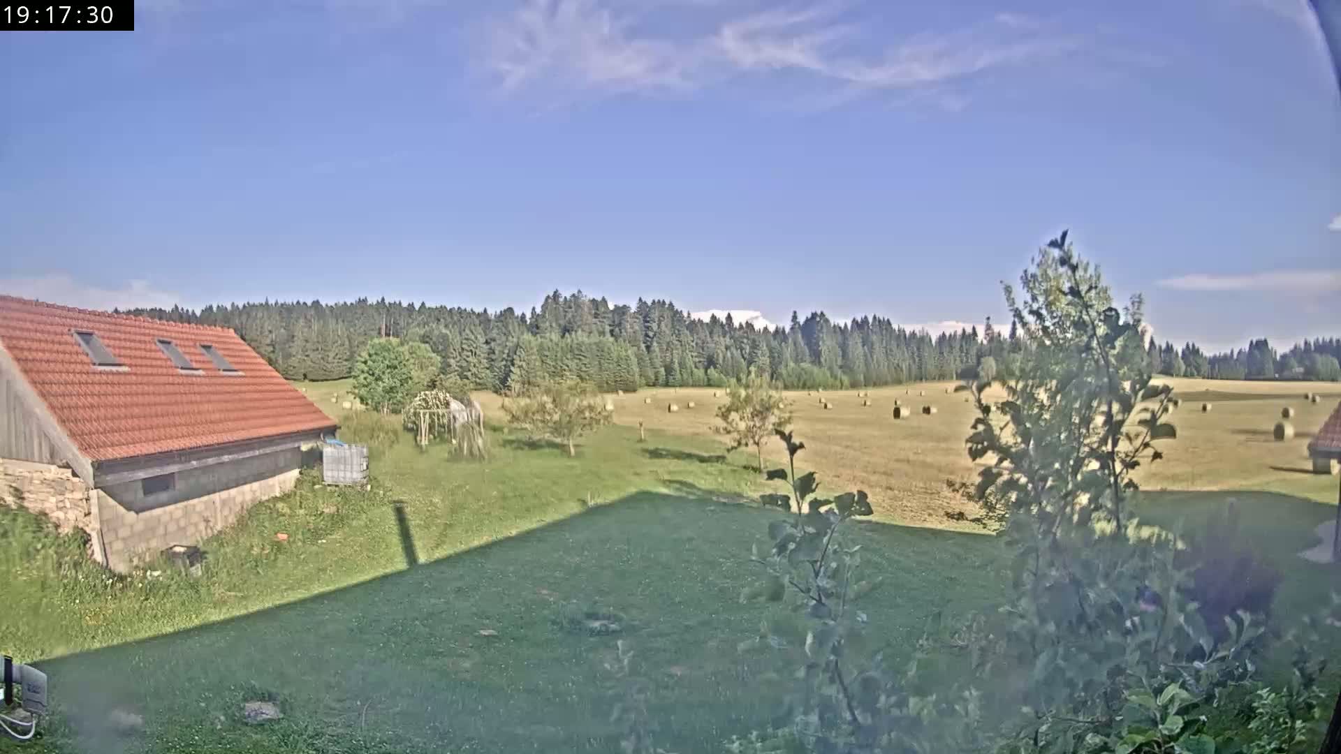 A red-roofed building sits near a grassy field with hay bales scattered across it, all under a mostly clear blue sky.