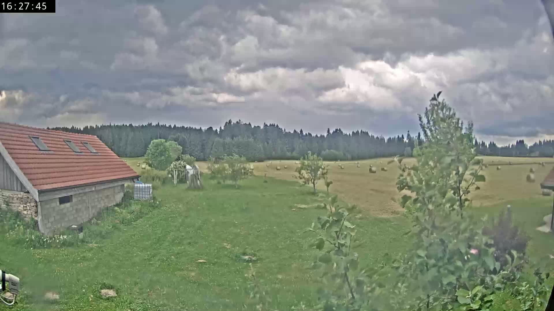 A red-roofed shed sits on a grassy yard overlooking a hay field dotted with hay bales, under a cloudy sky.