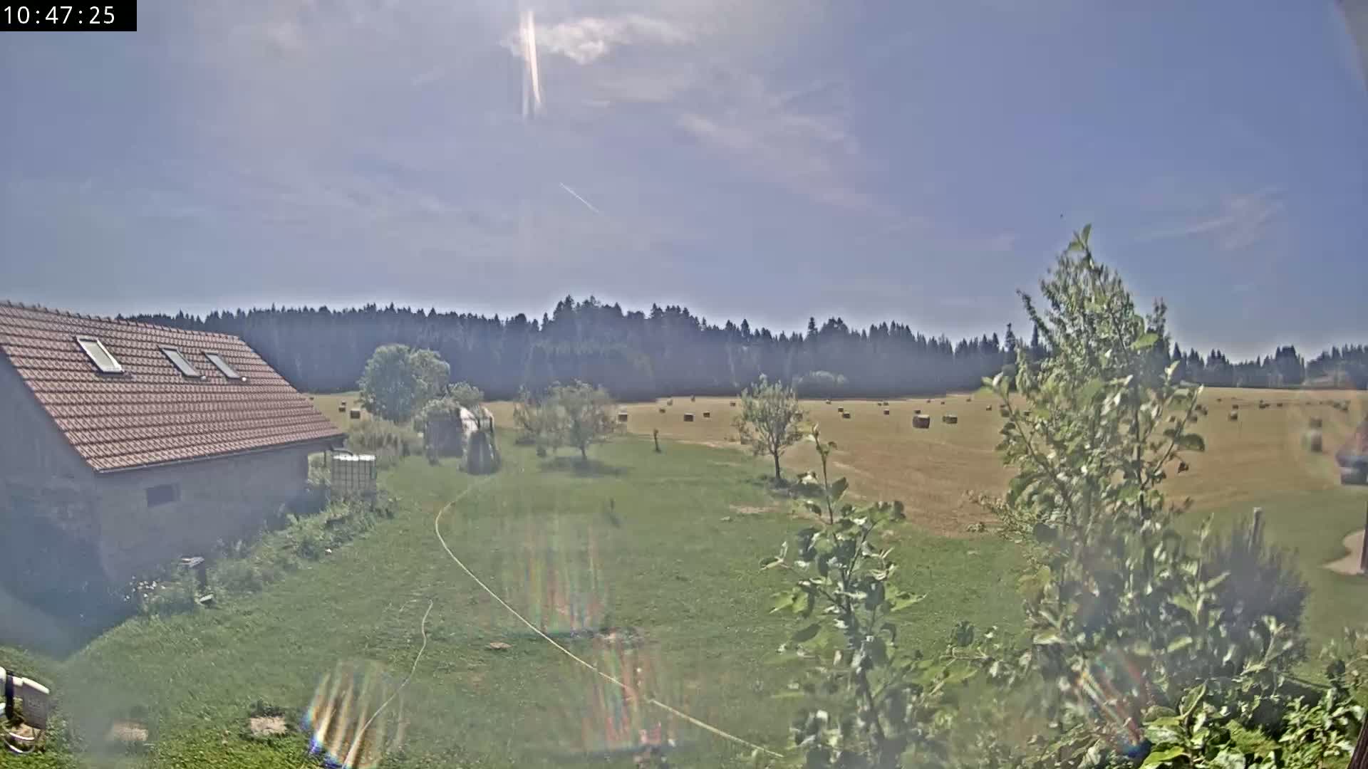 A brown-roofed building sits on a grassy yard next to a field of hay bales under a mostly sunny sky.