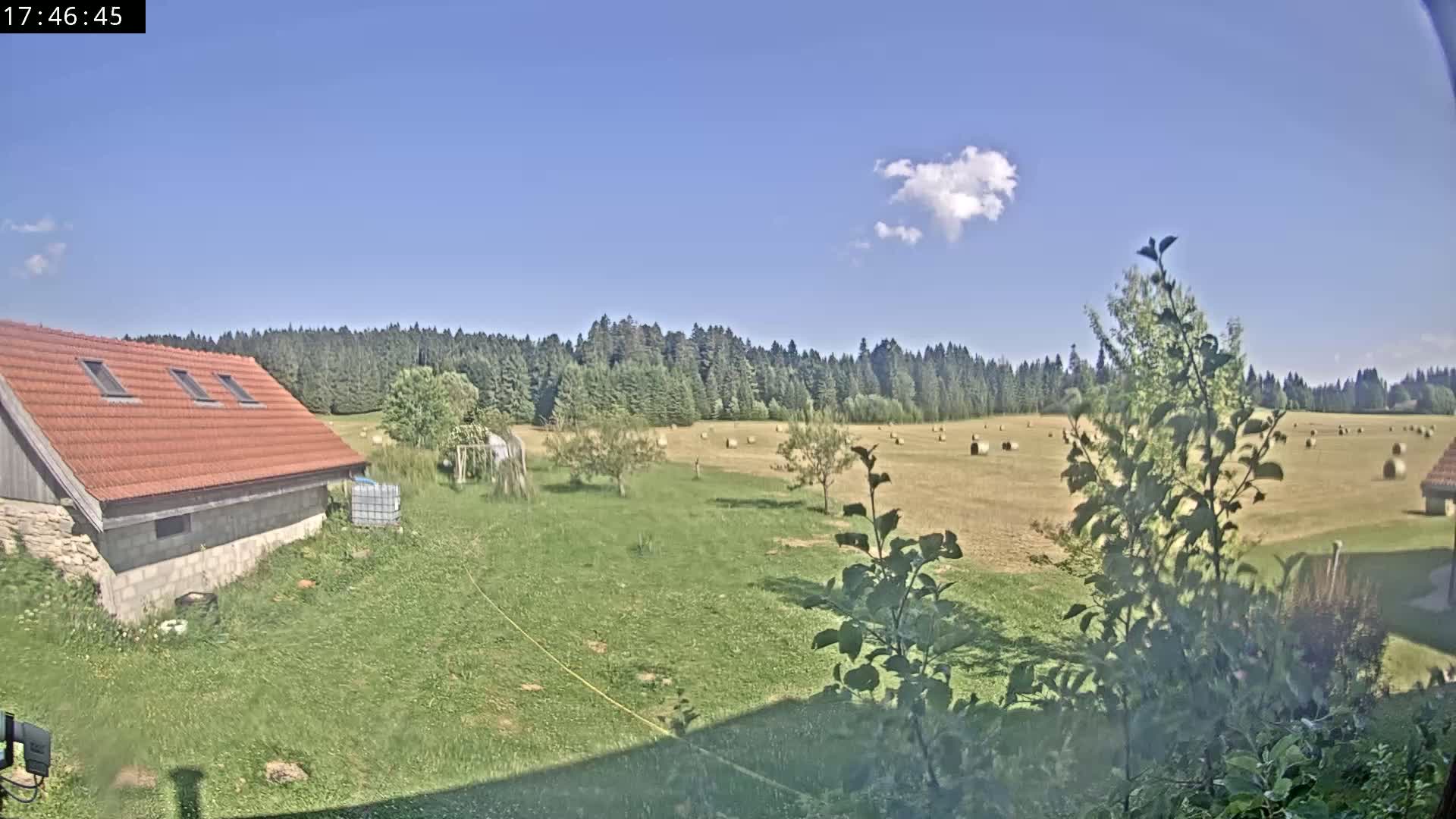 A red-roofed building sits on a grassy area next to a field of hay bales, under a mostly sunny sky.