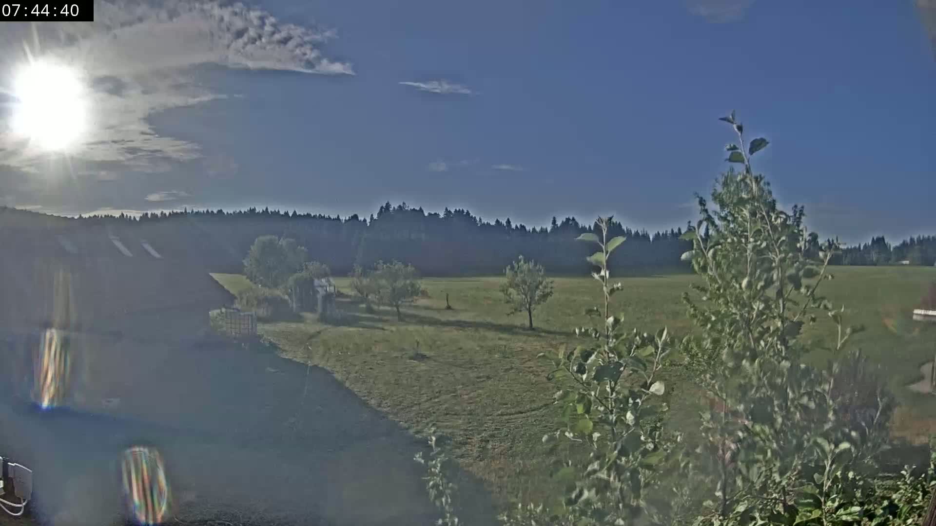 A partly sunny day reveals a grassy field with a few trees, a building, and a line of trees in the distance.