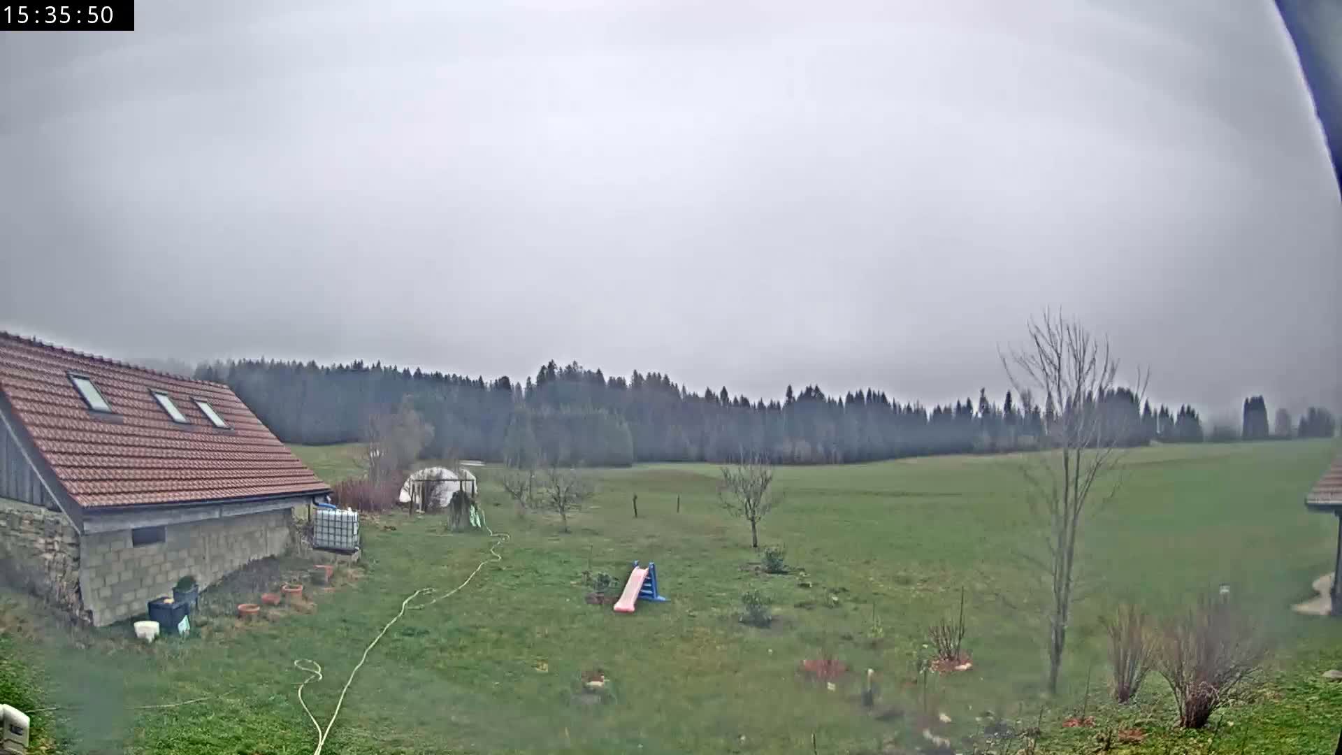 A vast, snow-covered winter landscape is depicted under a grey, overcast sky, showcasing a rustic building on the left, several bare trees, and a dense, snow-laden forest in the distance.