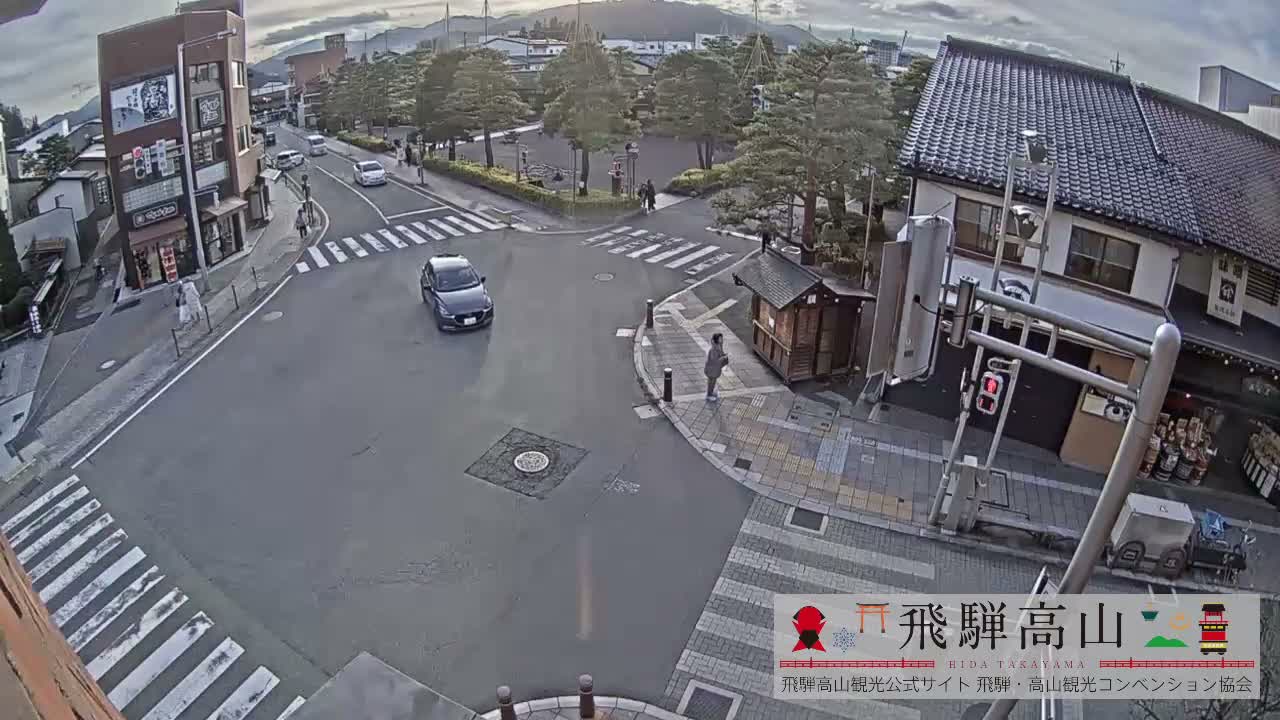 An elevated view shows a street intersection with vehicles and pedestrians, surrounded by traditional and modern buildings, trees, and distant mountains under a partly cloudy sky.