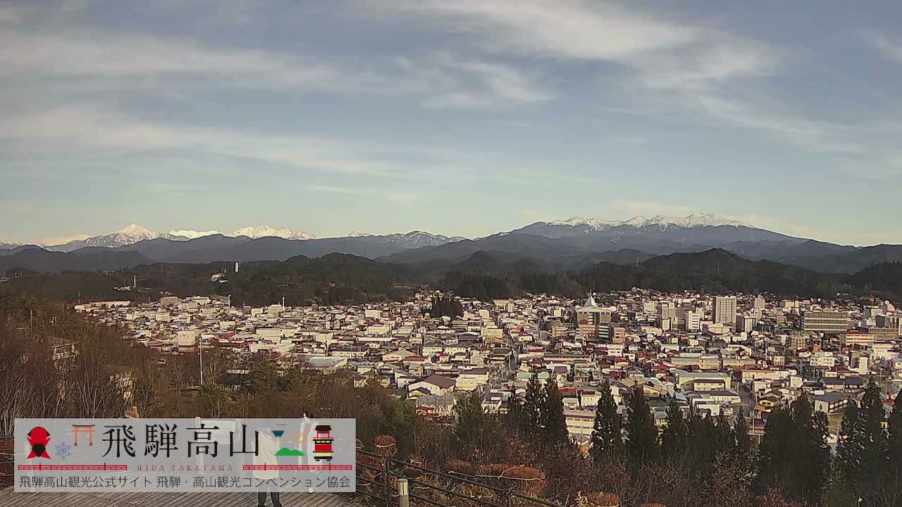 A sprawling town with dense buildings and red rooftops is nestled among wooded hills, backed by distant snow-capped mountains under a partly cloudy sky.