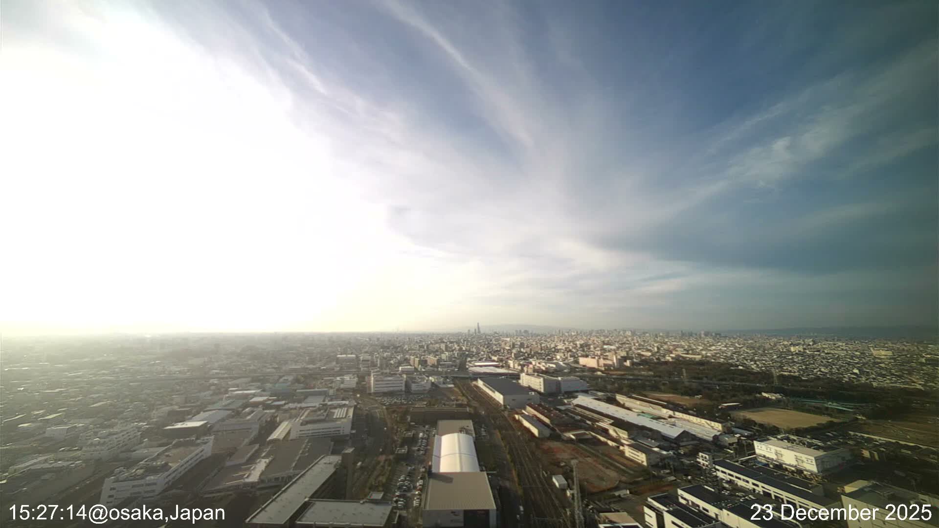 An expansive high-angle view captures a sprawling city under a partly cloudy sky with bright, hazy sunshine, showing numerous buildings and structures stretching towards a distant horizon.