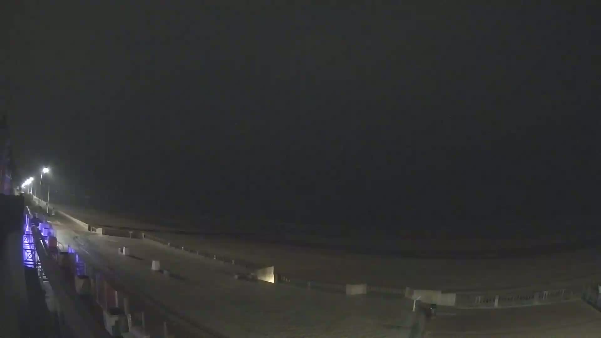 A nighttime, hazy view shows a beach and boardwalk extending into the distance under dimly lit streetlights.