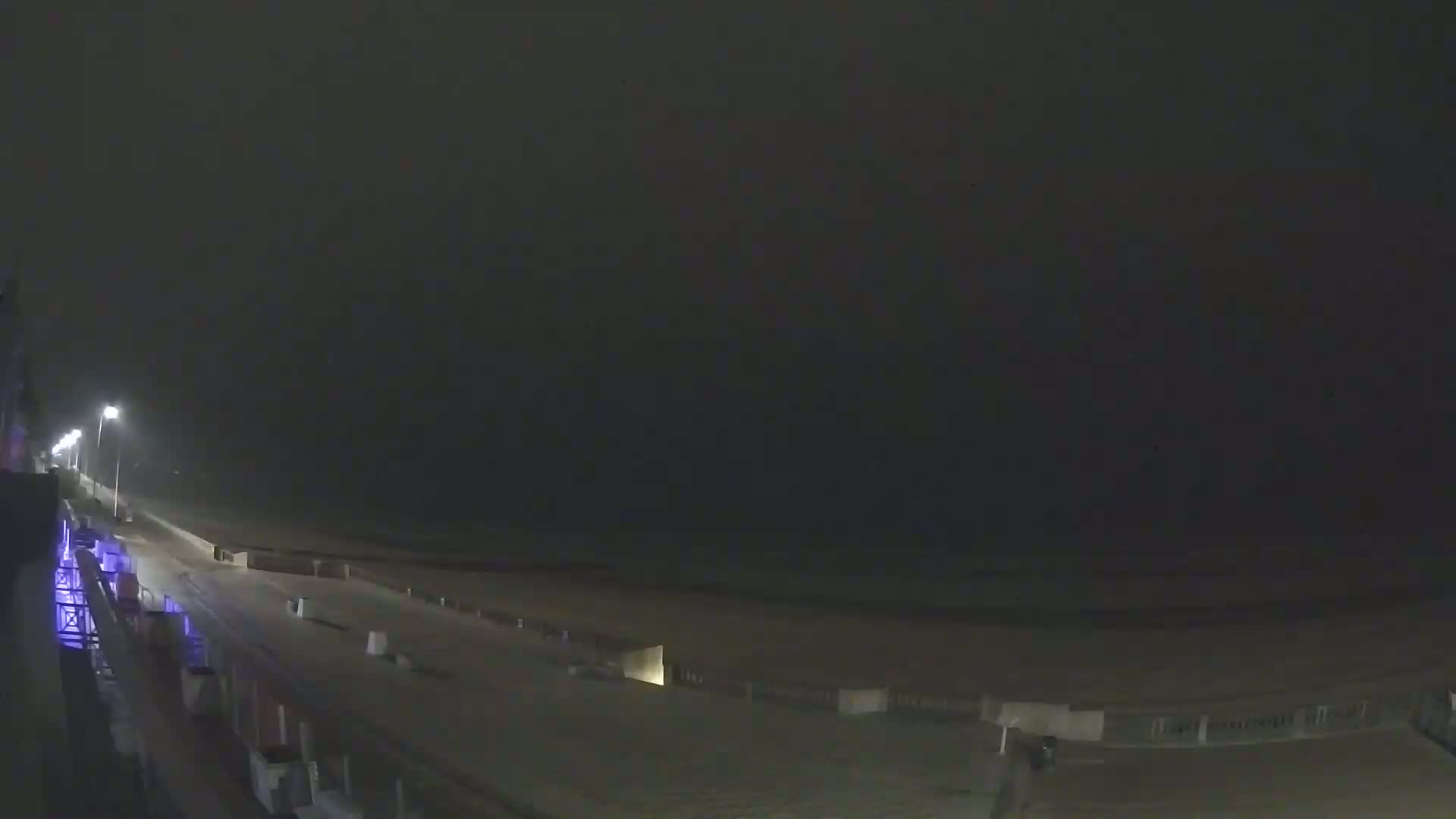 A dark, nighttime view of a mostly empty beach and boardwalk under a hazy sky.