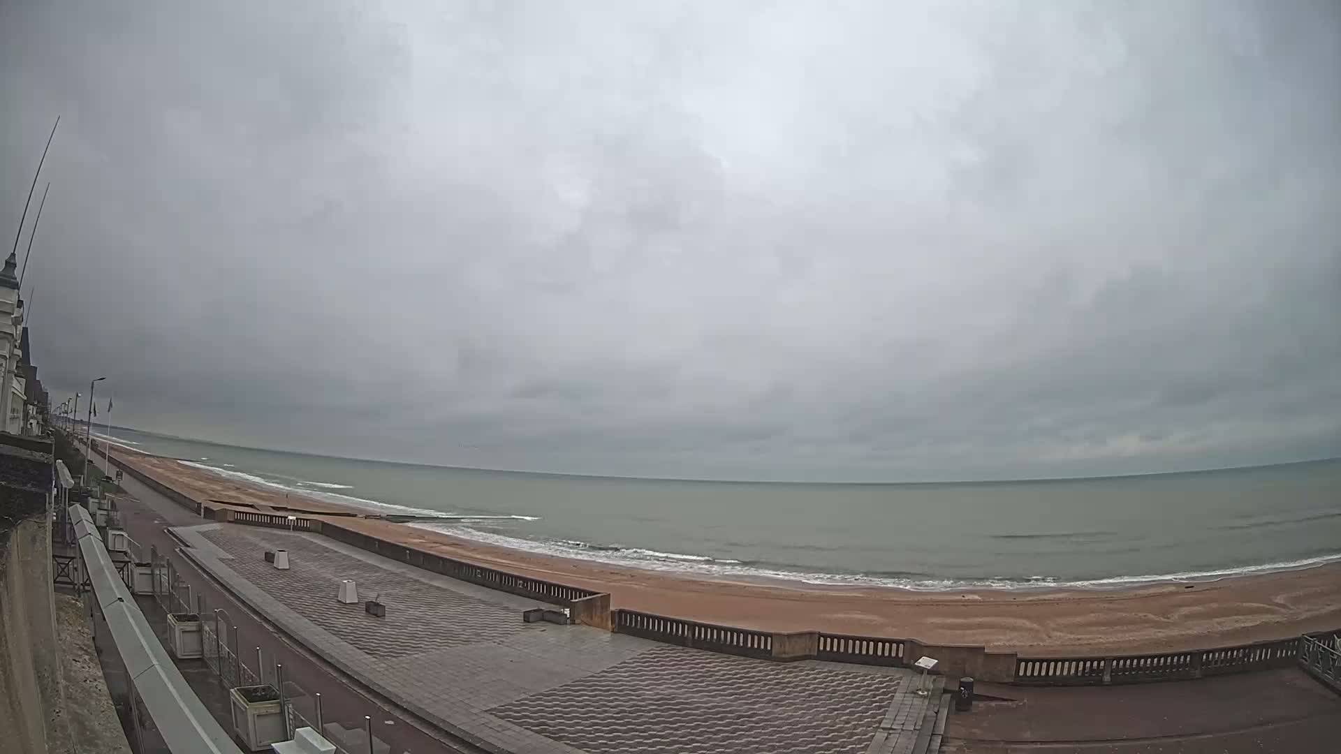A wide shot shows a mostly empty beach and promenade on an overcast day, with gentle waves lapping at the shore.