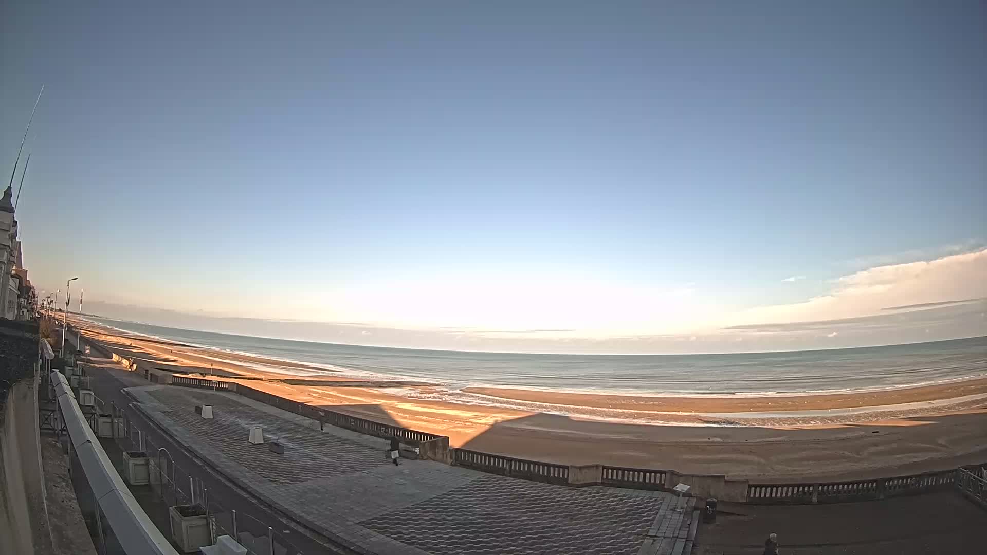 A mostly clear sky overlooks a wide sandy beach and calm ocean, with a paved walkway and building visible in the foreground.