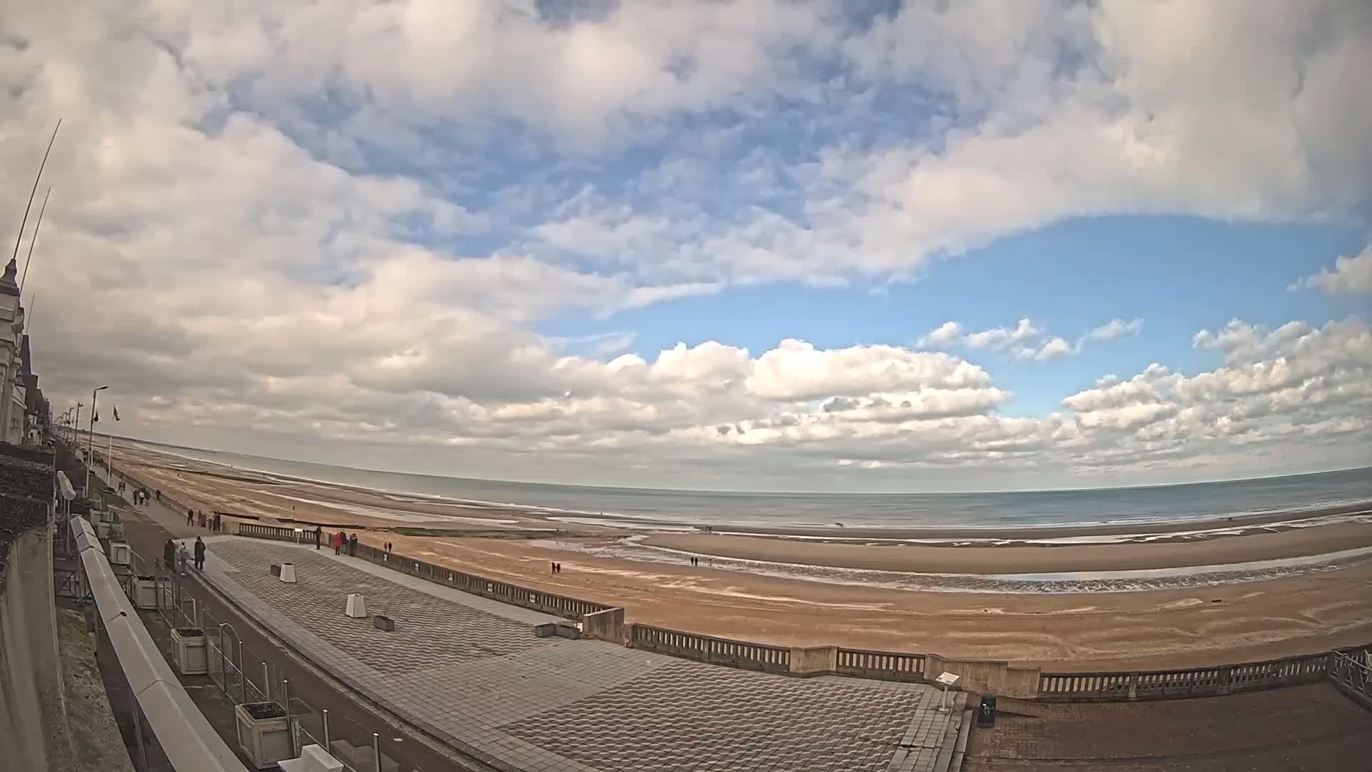 A wide, partly cloudy sky overlooks a long, sandy beach with a few people walking along a paved promenade next to a building.