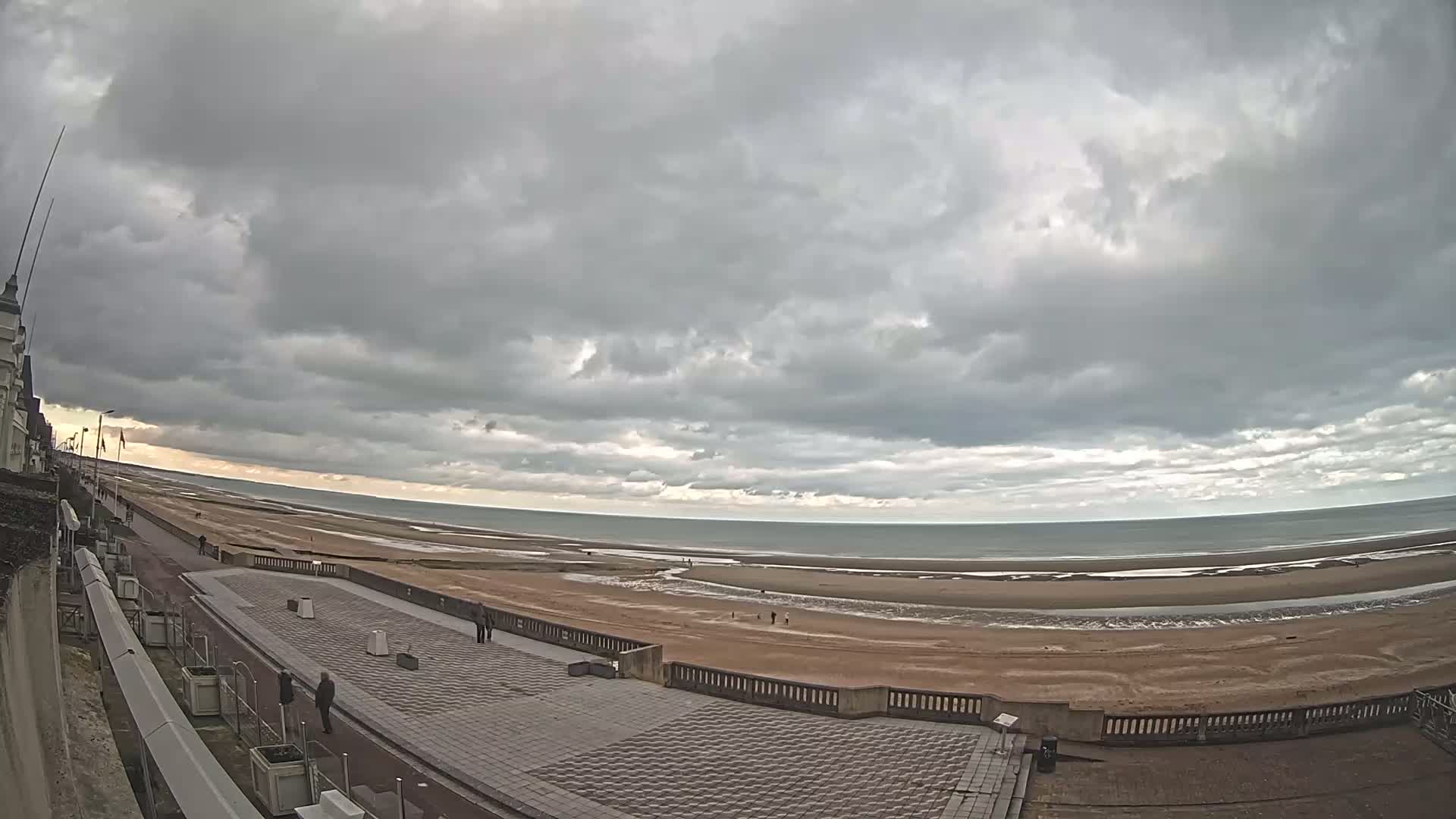 A wide shot of a beach with a paved walkway and a building in the distance under an overcast sky.