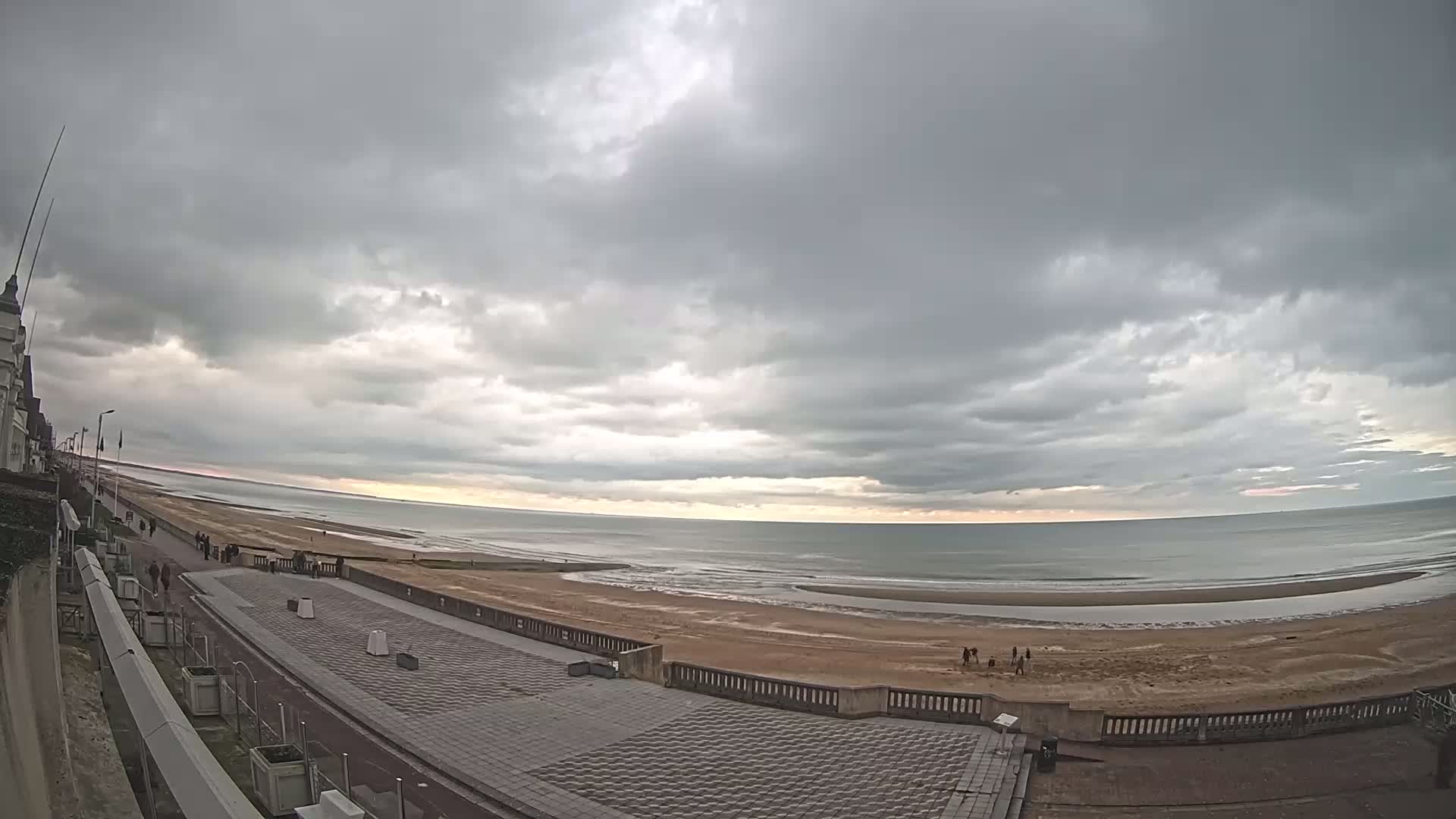 A mostly cloudy sky overlooks a wide, sandy beach and calm ocean with a paved walkway and building in the foreground.