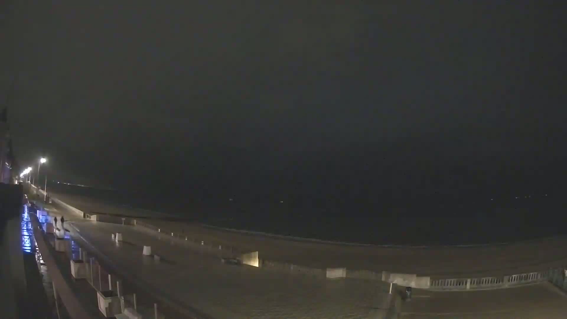 A dark, rainy night scene shows a wet boardwalk overlooking a beach and ocean under a mostly overcast sky.