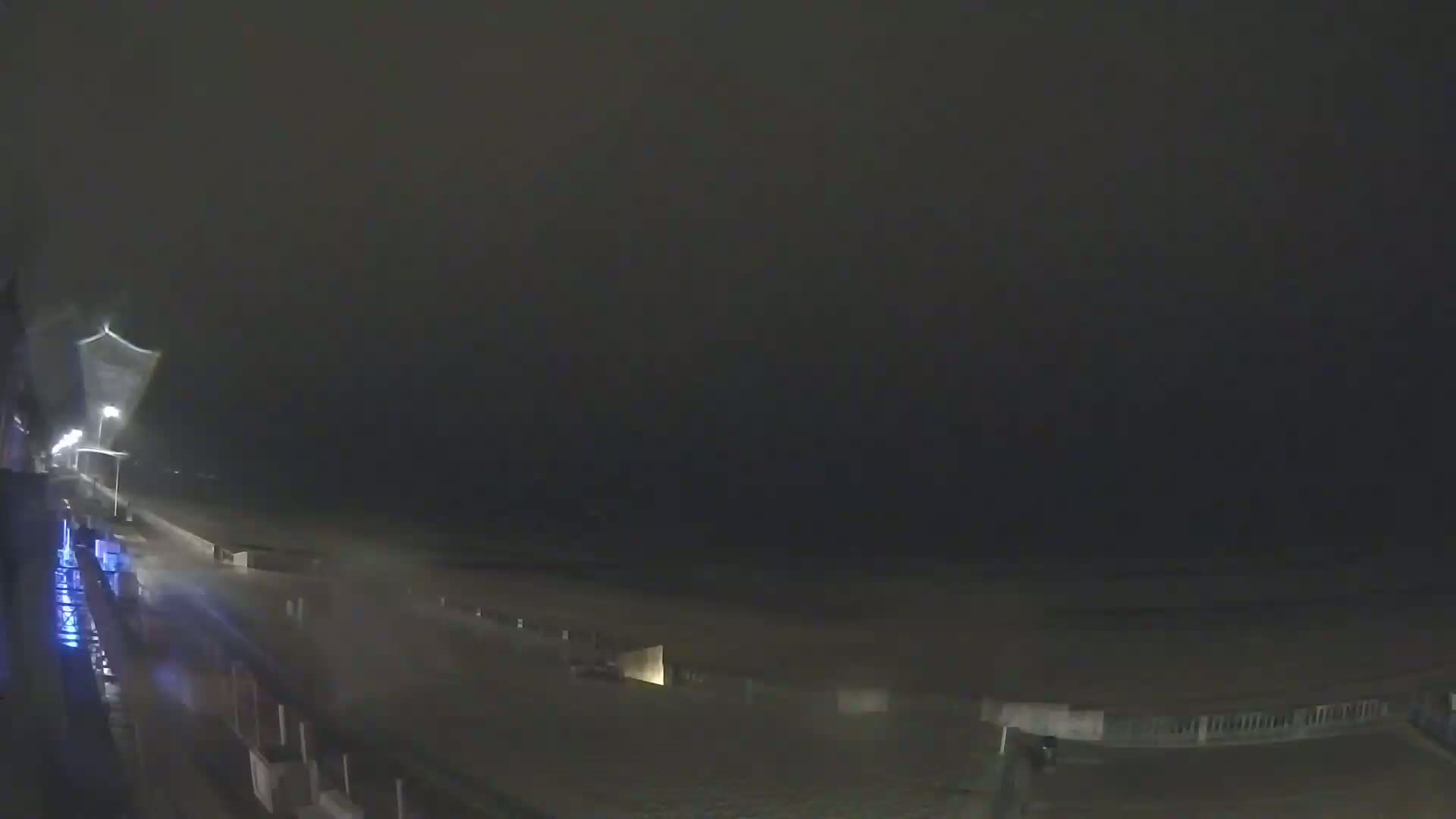 A nighttime coastal scene shows rain falling on a dark beach and promenade under a cloudy sky.