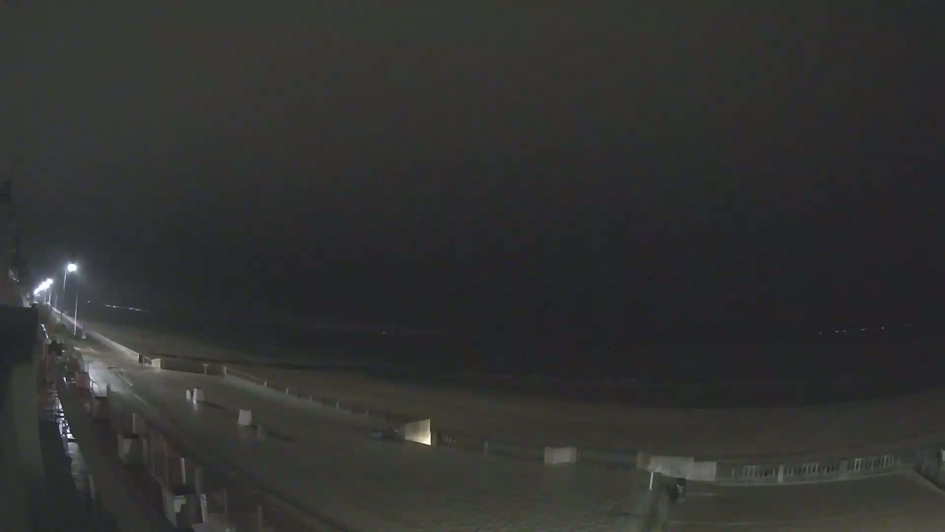 A dark, nighttime view of a wet beach road leading to a dark ocean under an overcast sky.