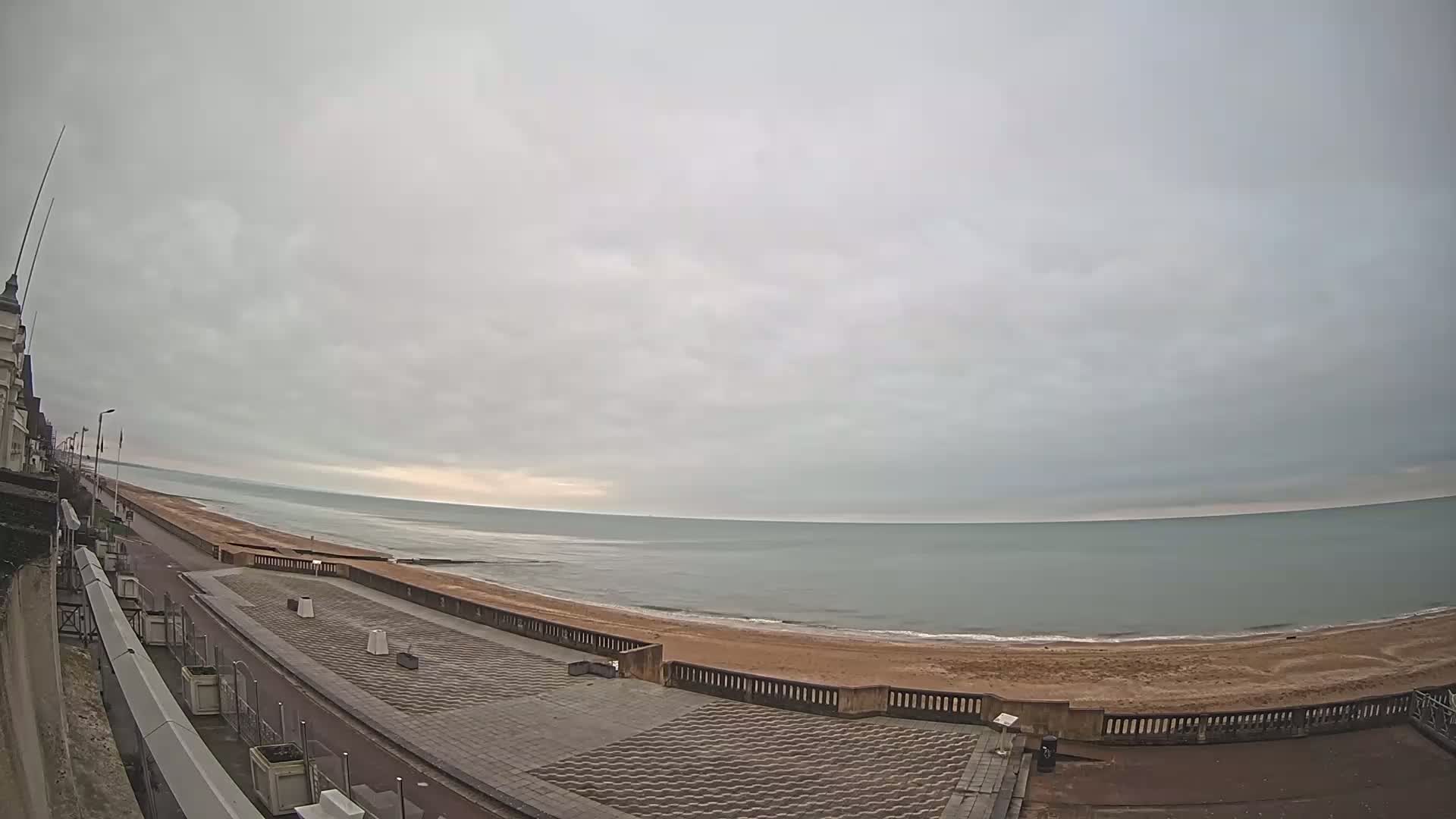 A mostly cloudy day shows a calm sea with a sandy beach and a paved promenade next to a building.