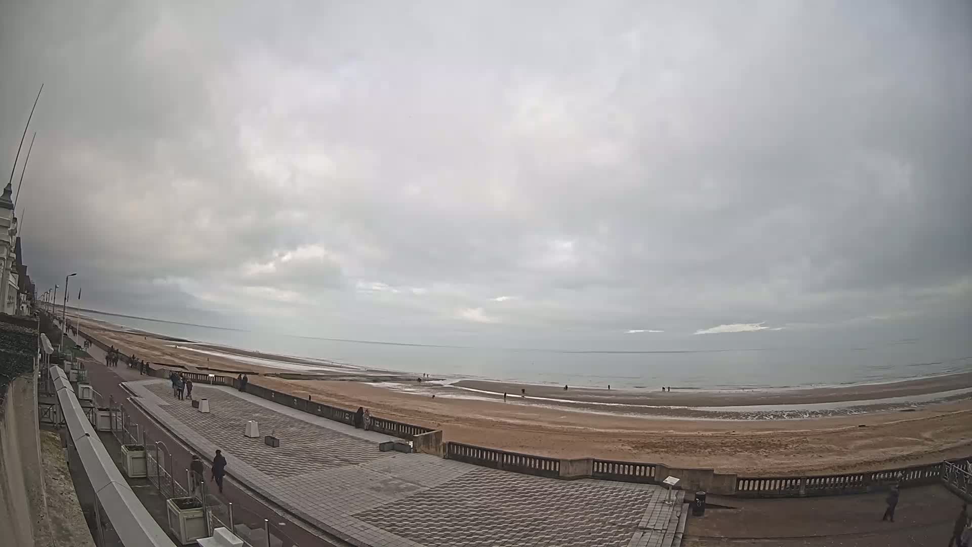 A wide shot shows a mostly overcast sky over a beach with a paved walkway and a few people walking along it.