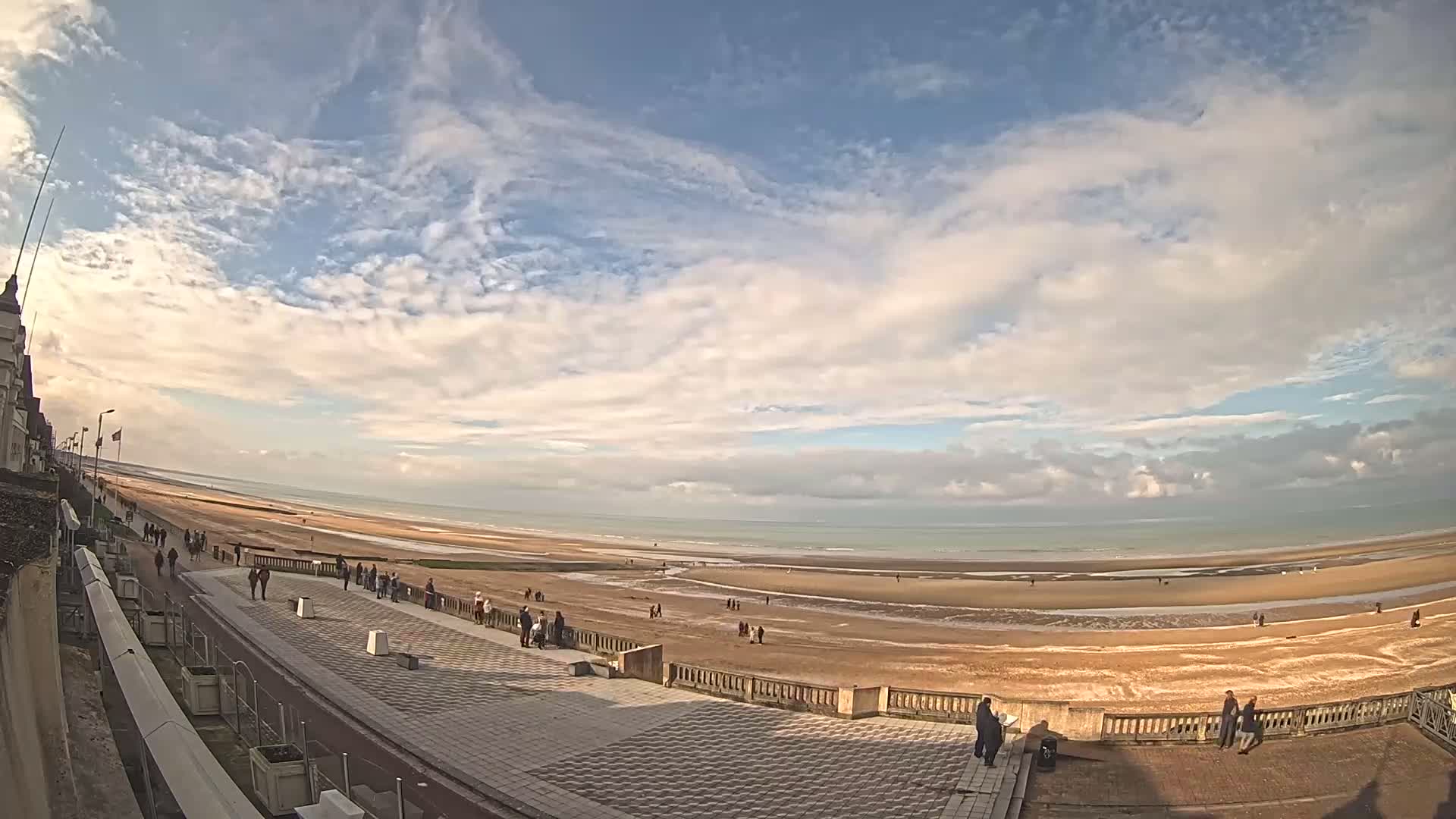 A wide shot shows a sandy beach stretching to a calm ocean under a partly cloudy sky, with people walking along a paved promenade next to the beach.