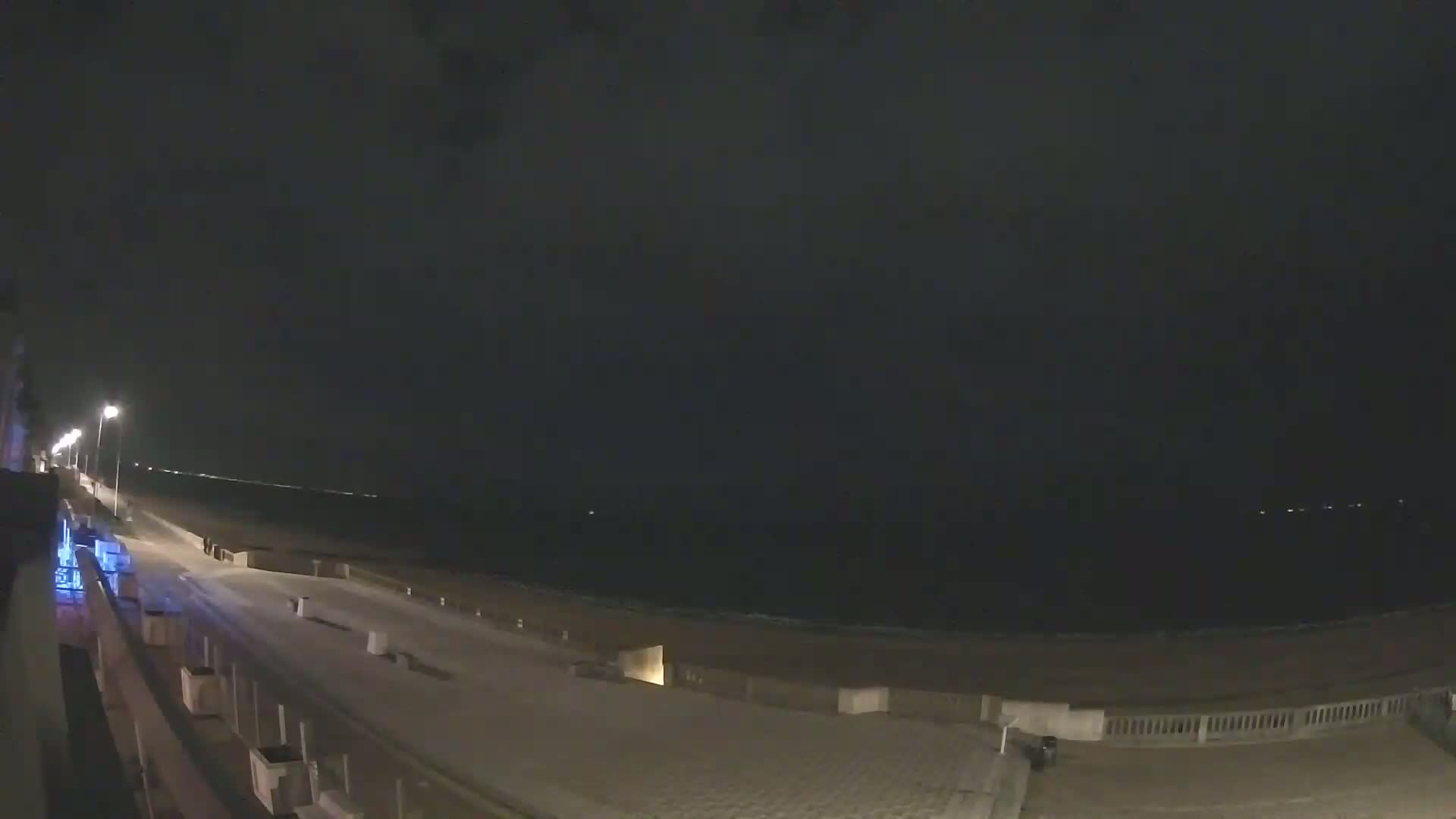 A nighttime view of a mostly empty boardwalk and beach, lit by streetlights under a dark sky.