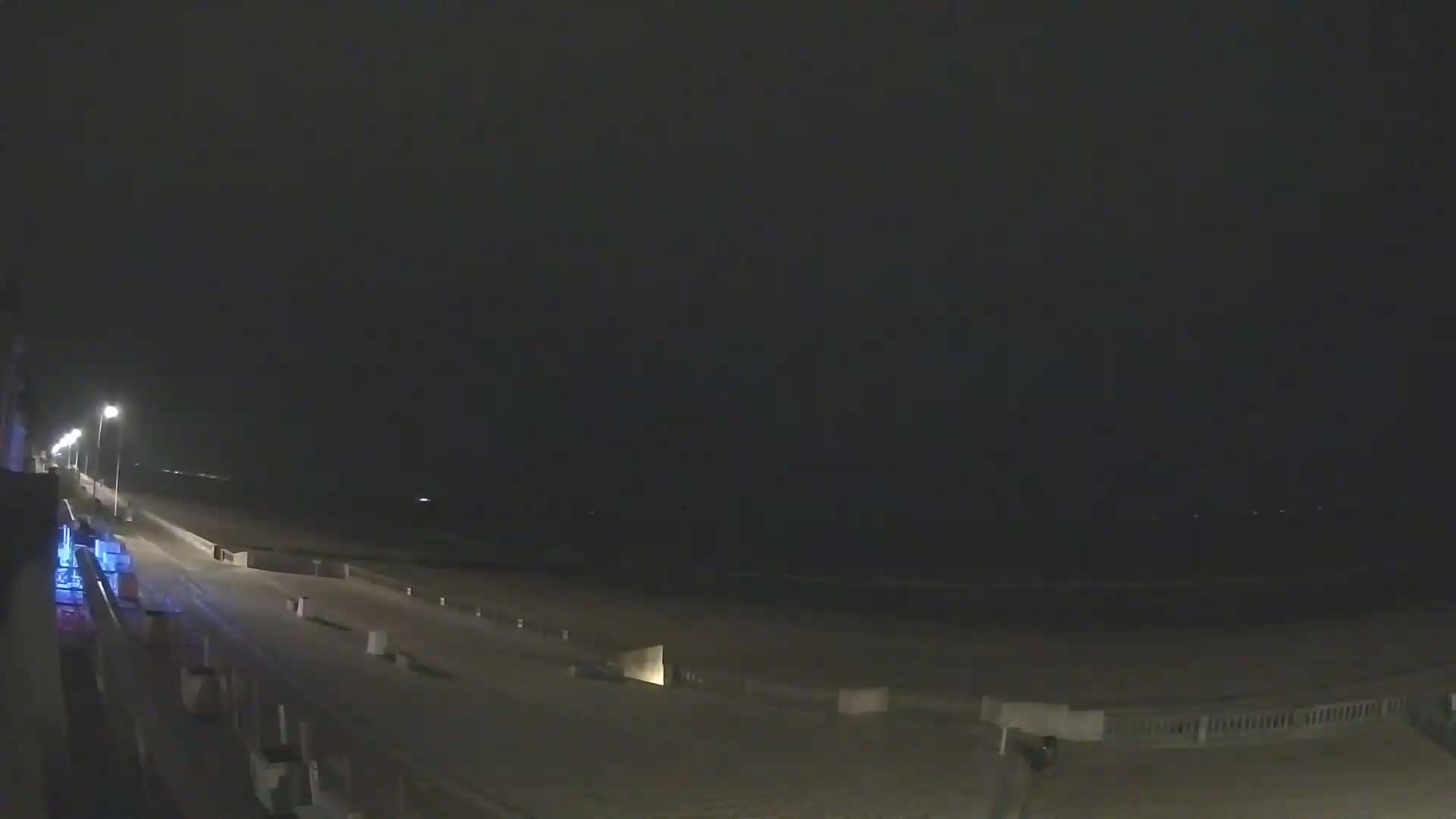 A nighttime view of a mostly empty beach boardwalk with a few lights illuminating the scene under a dark sky.