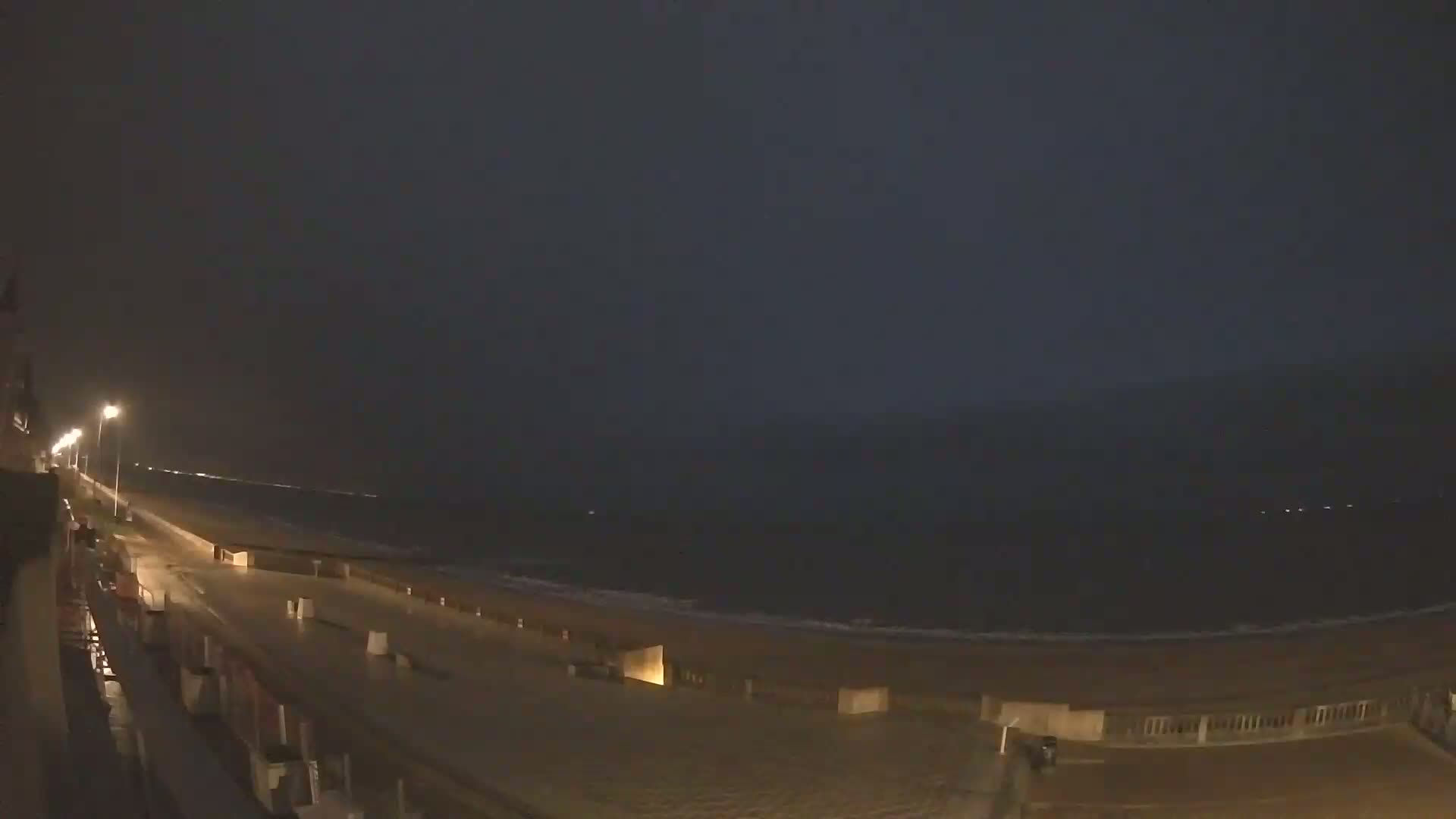 A rain-soaked boardwalk stretches alongside a dark, nighttime ocean under a stormy sky.