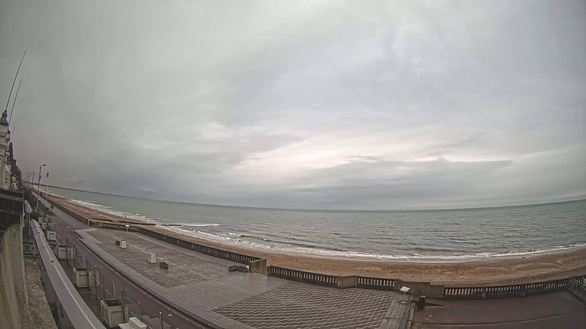 A mostly cloudy day shows a wide sandy beach and calm ocean waves, viewed from a paved walkway beside a building.