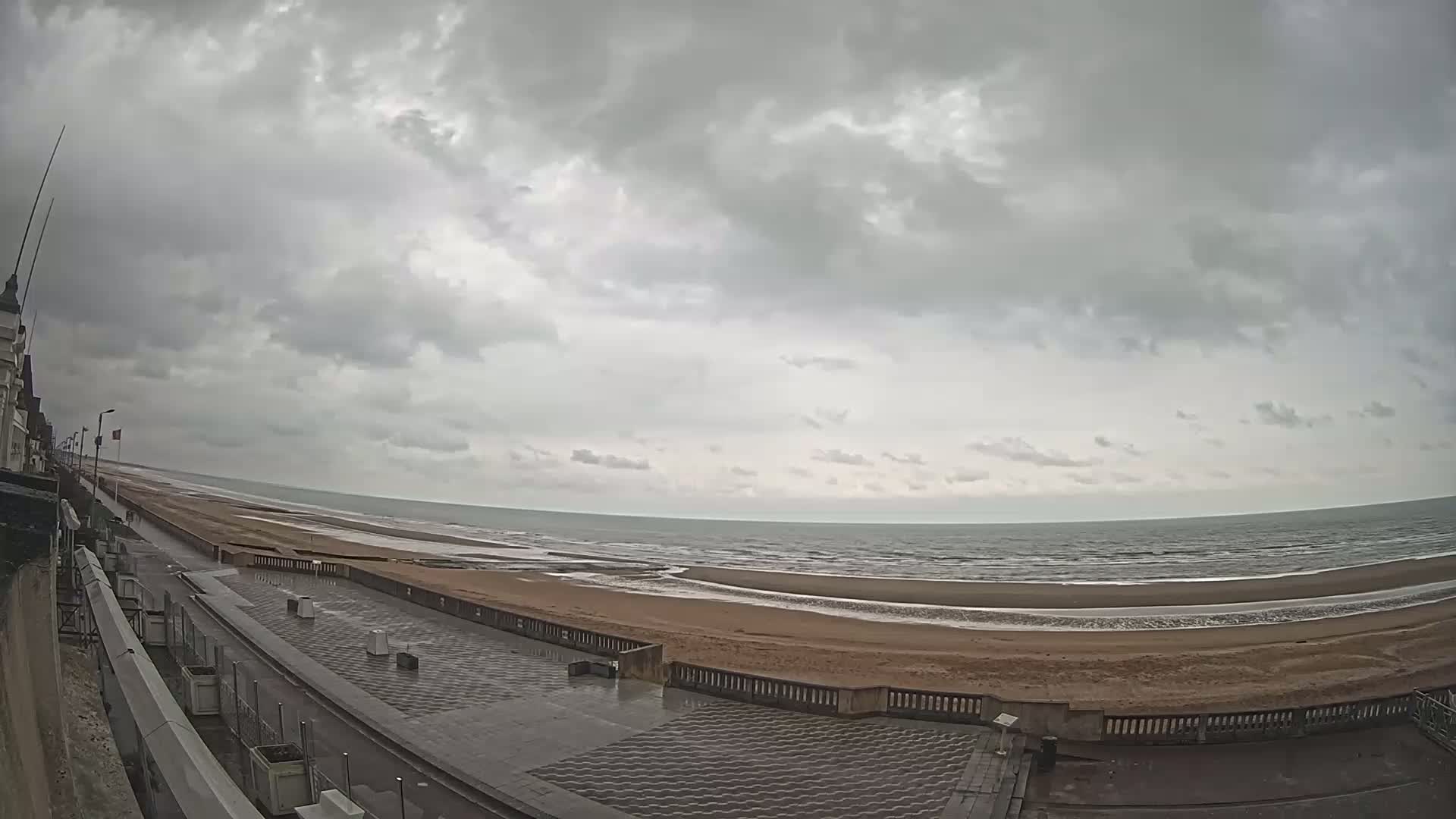 A wide shot shows a mostly cloudy sky over a long, sandy beach and calm ocean, with a paved walkway and building visible in the foreground.