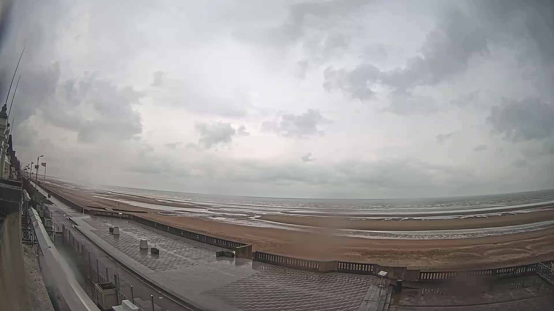 A wet, paved walkway next to a wide, sandy beach and ocean under a cloudy sky.