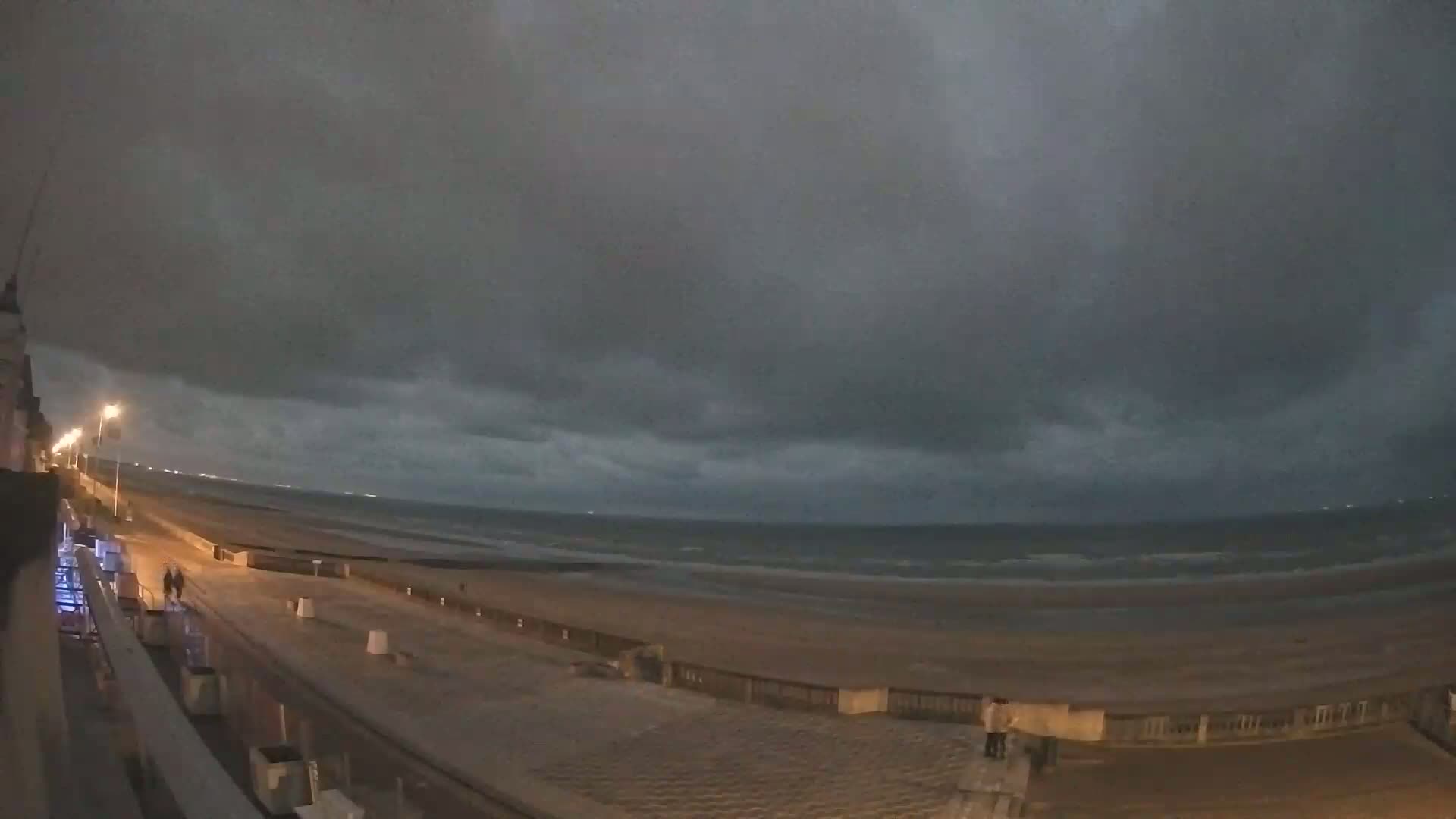 A nighttime view of a beach with a dark, cloudy sky and a few people walking along the promenade.