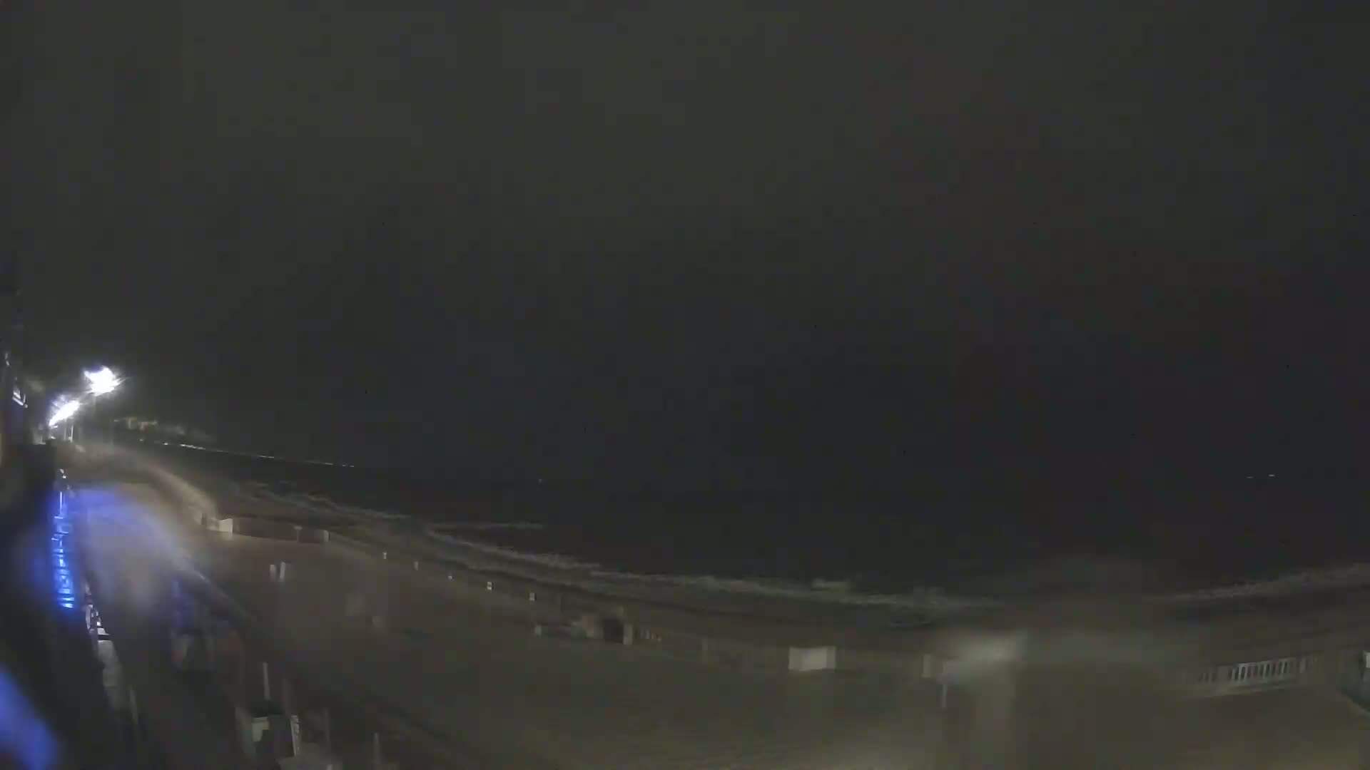 A nighttime view of a rain-swept beach with waves crashing on the shore, illuminated by streetlights.