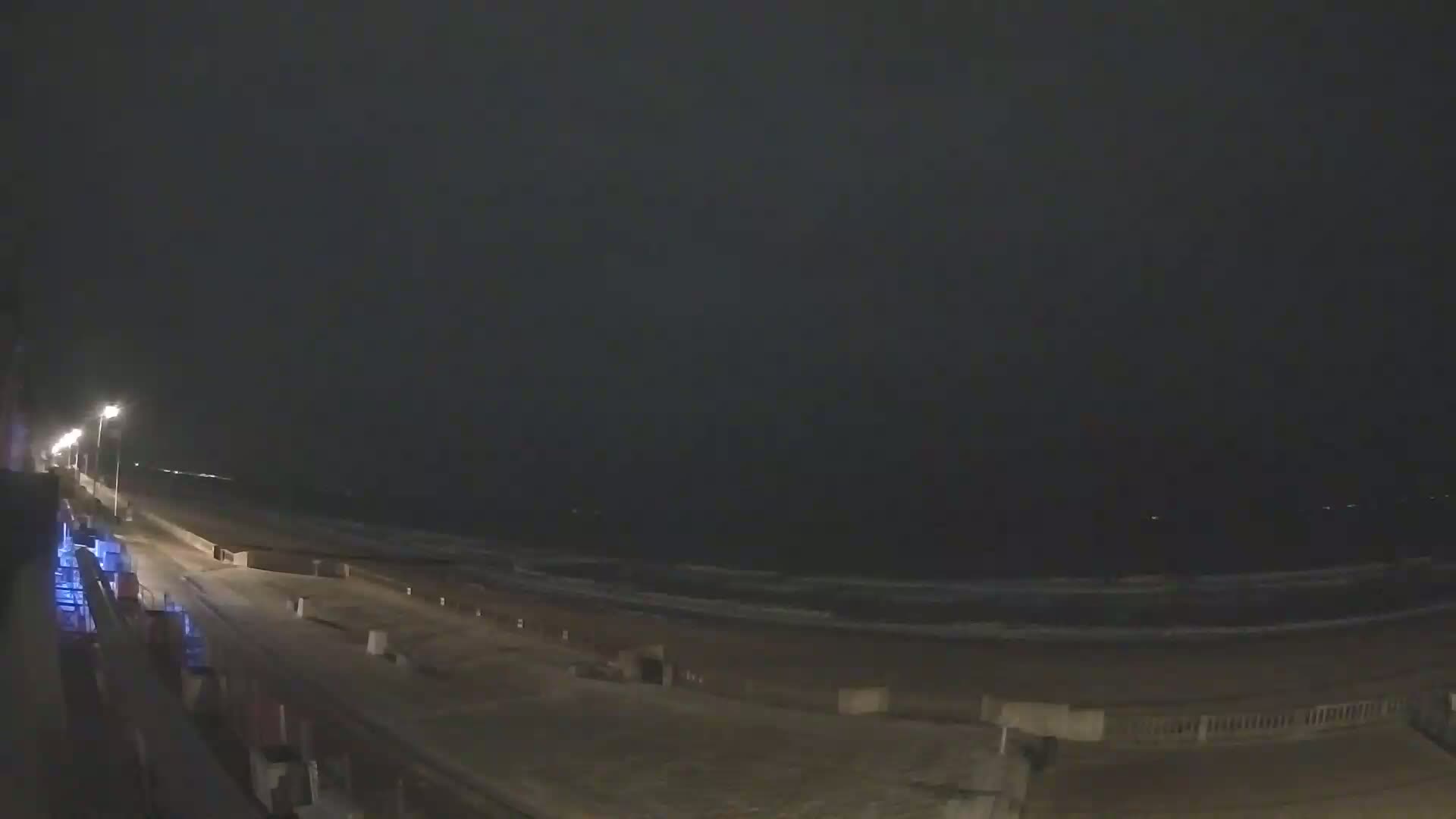 A nighttime view of a beach with dark, overcast skies, showing a boardwalk and gently lapping waves.