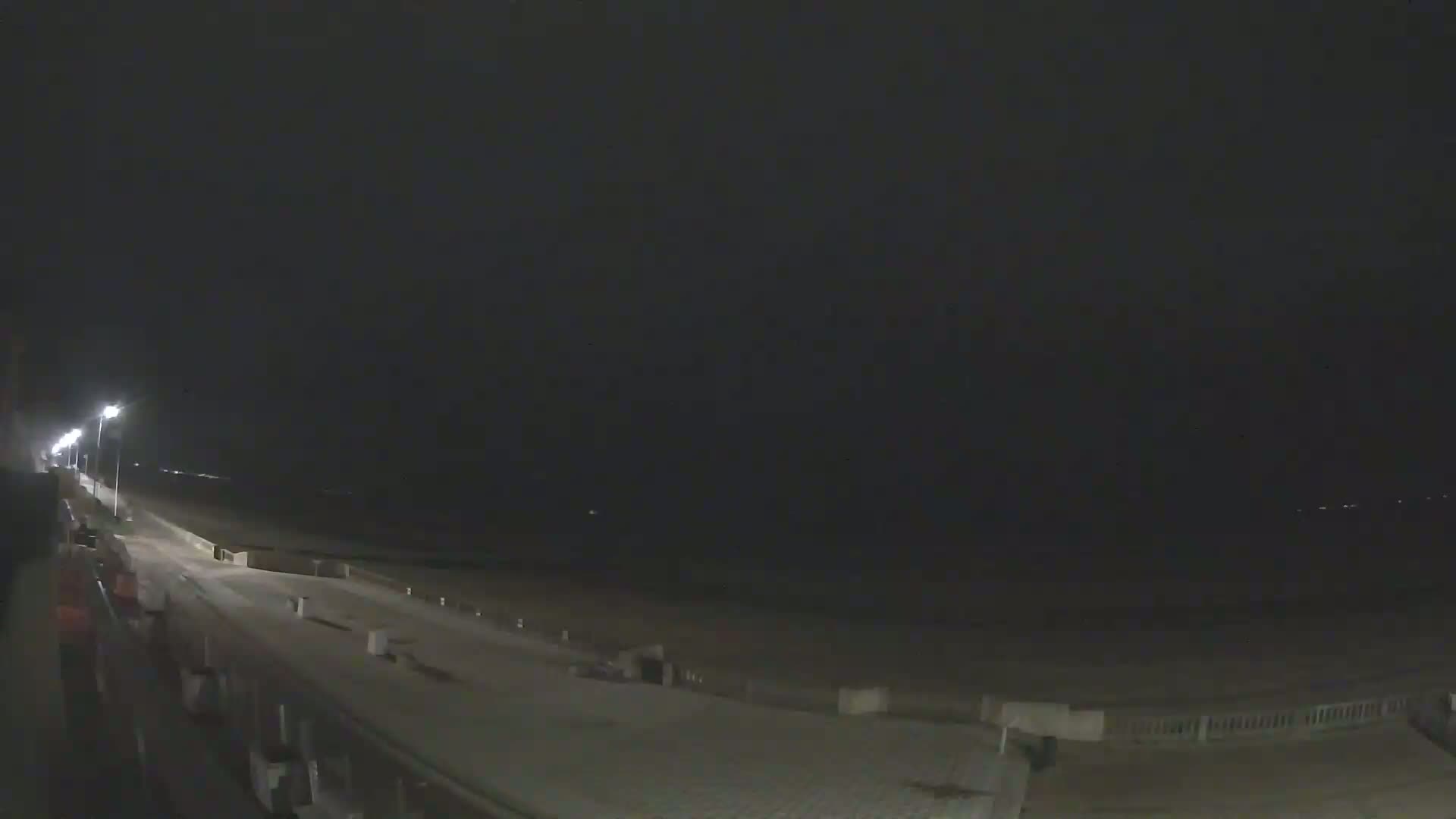 A dark, nighttime view of a mostly empty boardwalk next to a beach, illuminated by streetlights.