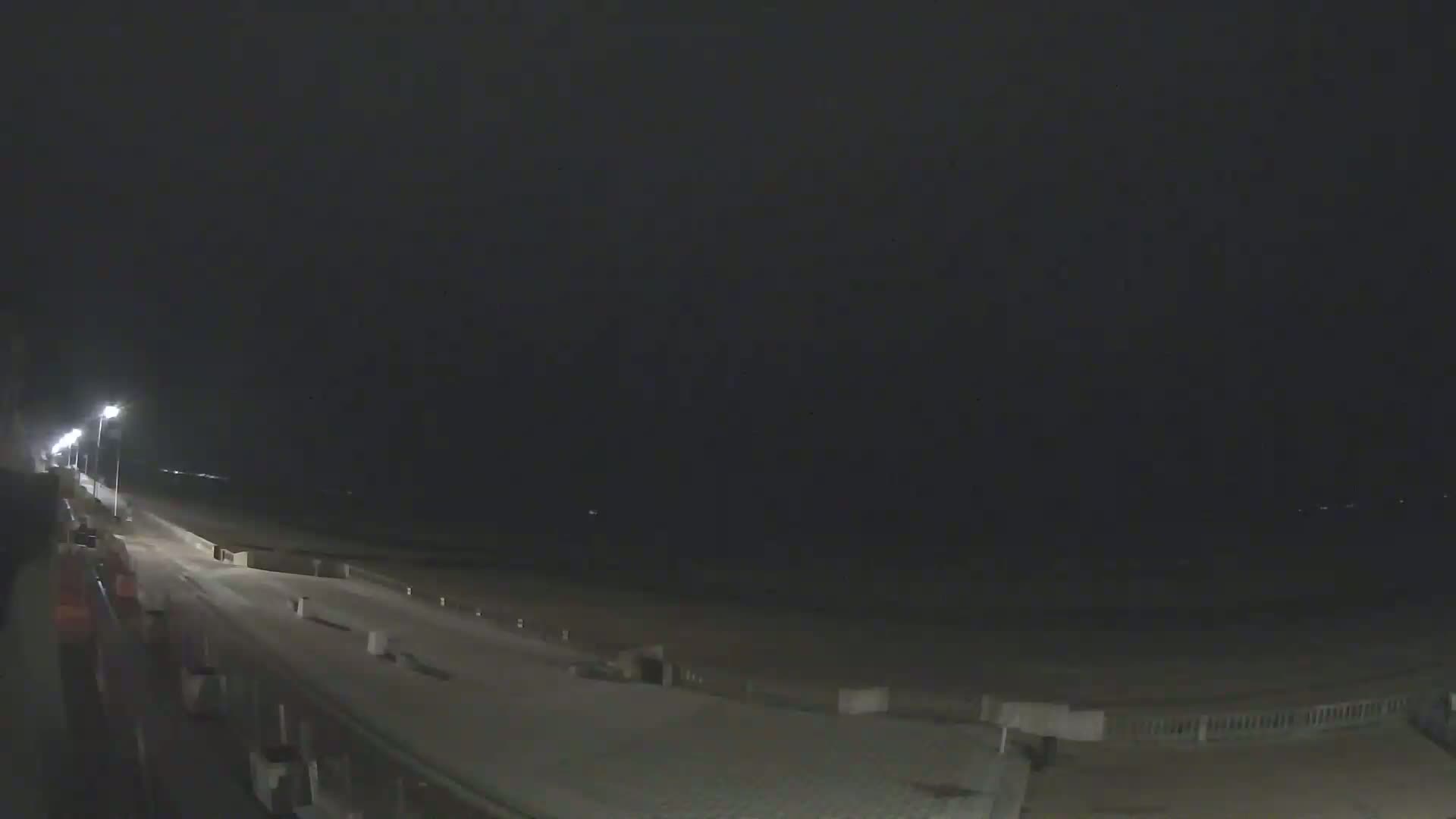A dark, nighttime view of a beach boardwalk with streetlights illuminating the path and a dark ocean in the background.