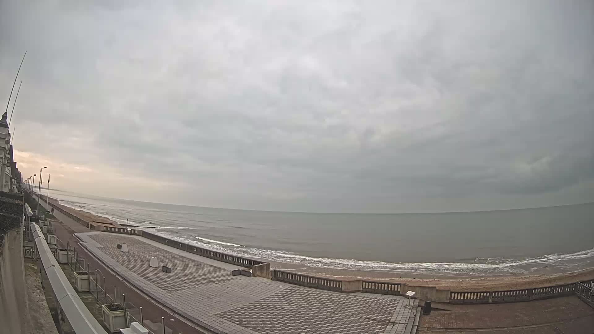 A paved seaside promenade overlooks a calm, grey ocean under an overcast sky.