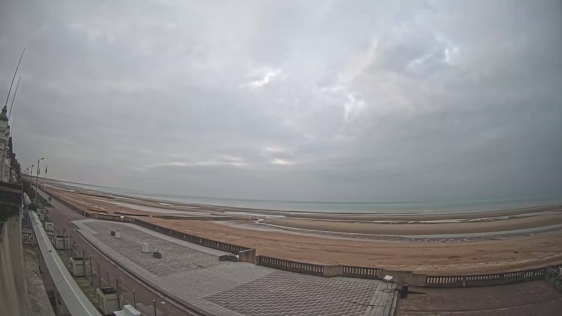 A wide, overcast sky overlooks a paved seaside promenade next to a wide, sandy beach at low tide, with the ocean visible in the distance.