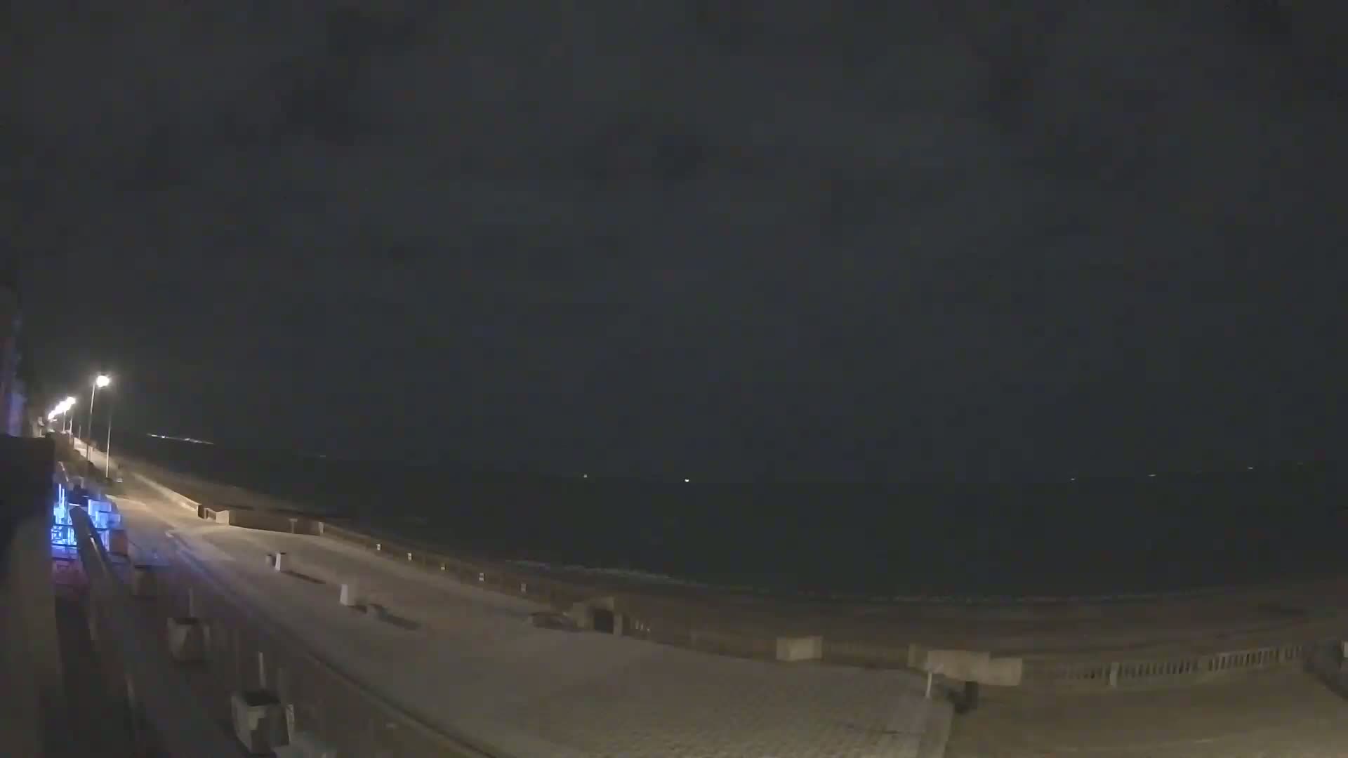 A nighttime view shows a dark ocean under a cloudy sky, a paved walkway lined with lights leading to the water's edge.
