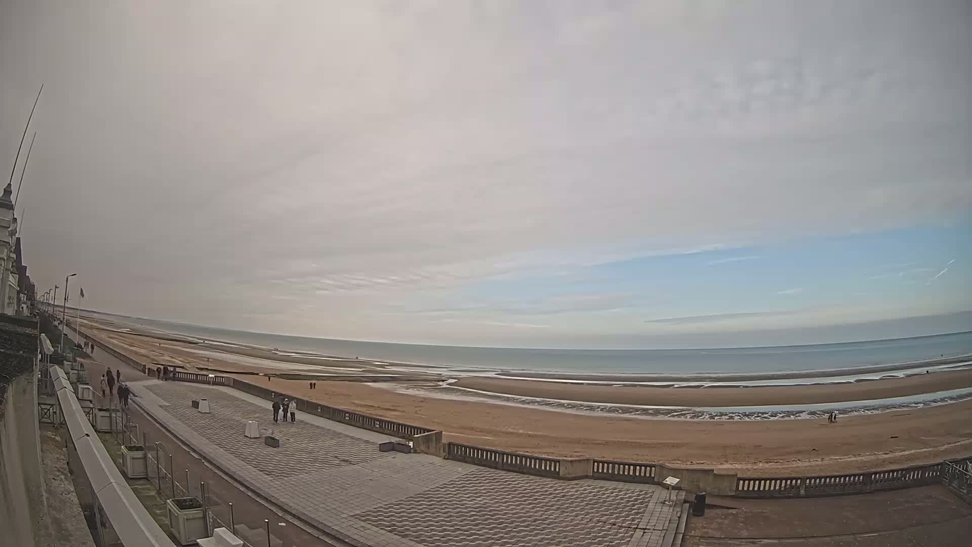 A wide, sandy beach stretches along a calm sea under a mostly cloudy sky, with a paved walkway and a few people visible near the shoreline.