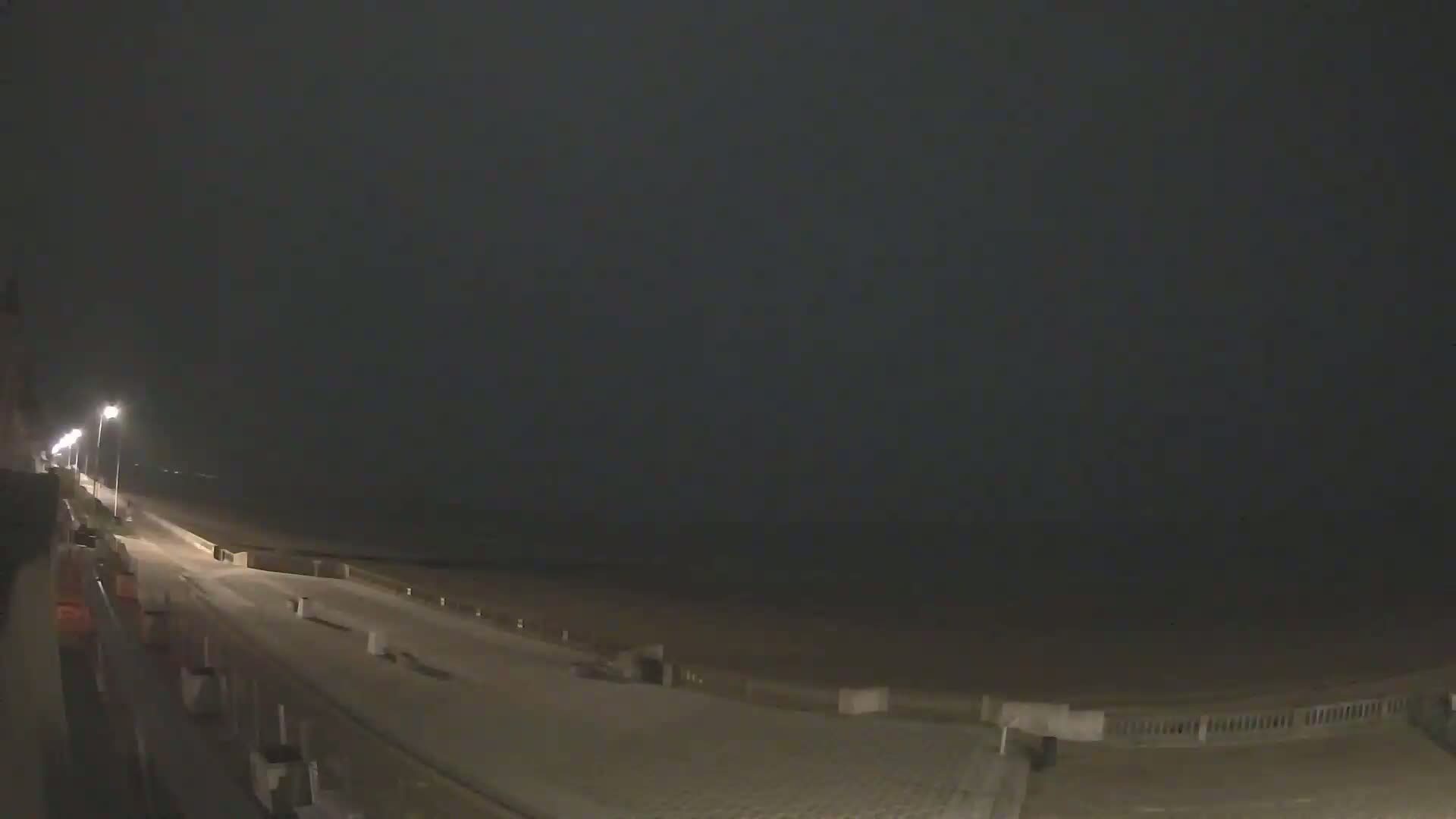 A nighttime view of a dimly lit beach promenade extends toward a dark, overcast ocean.