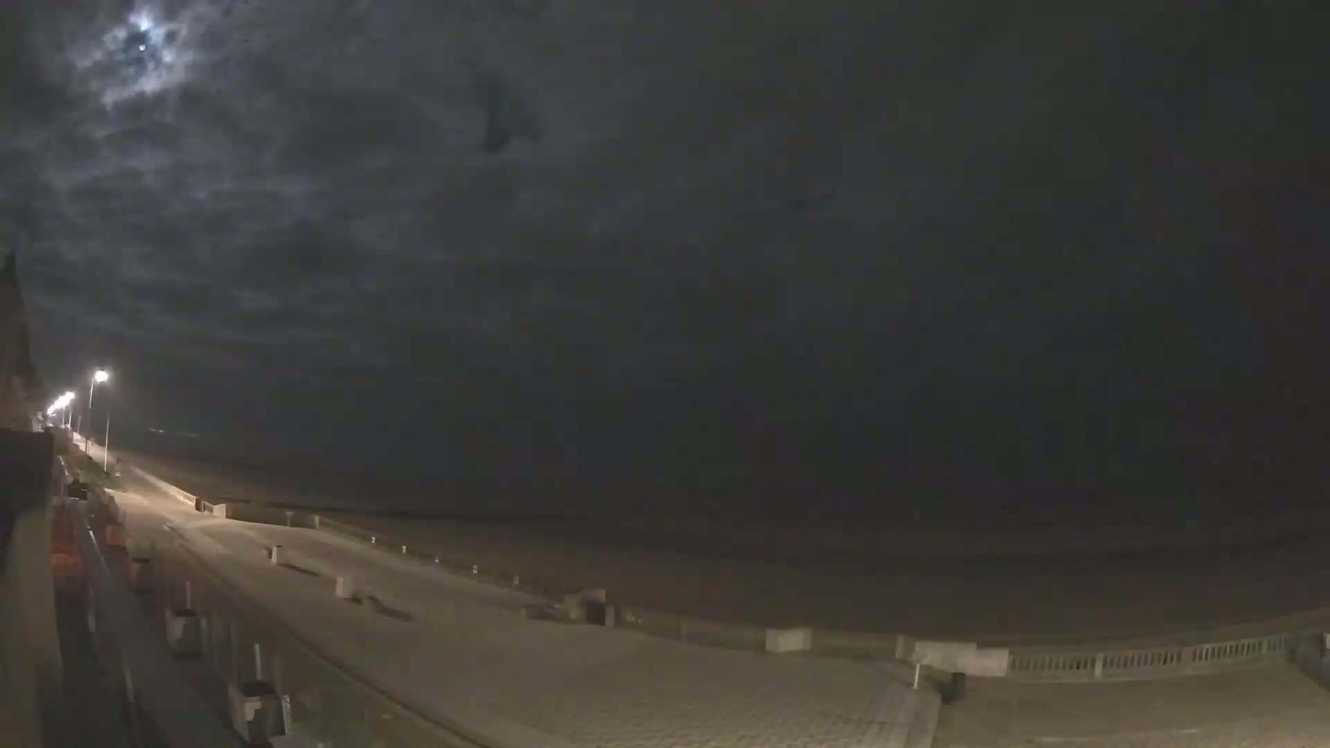 A moonlit night scene shows a paved walkway beside a beach, illuminated by streetlights under a cloudy sky.