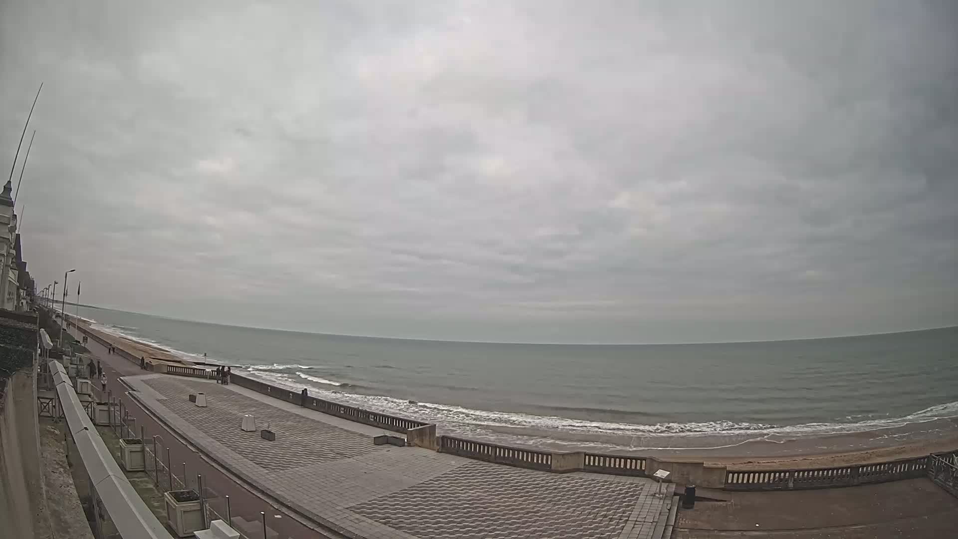 A paved seaside promenade with a few people walking along it, overlooks a beach with gentle waves under an overcast sky.