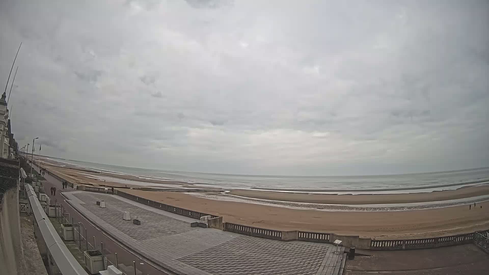 A wide shot shows a paved beachfront promenade overlooking a wide sandy beach and calm ocean under an overcast sky.