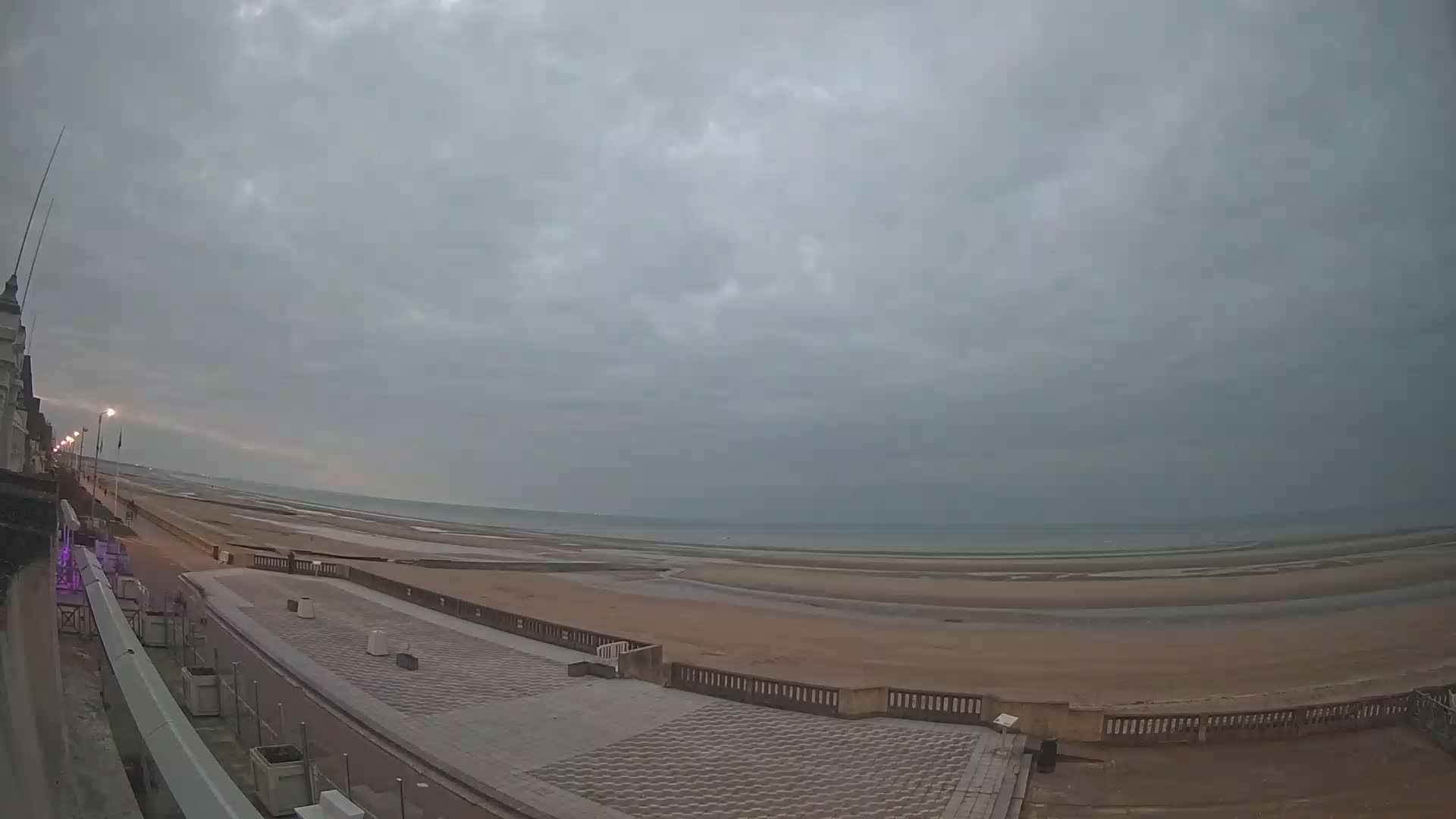A wide shot of a paved beachfront promenade overlooking a wide expanse of sandy beach and calm sea under an overcast sky.