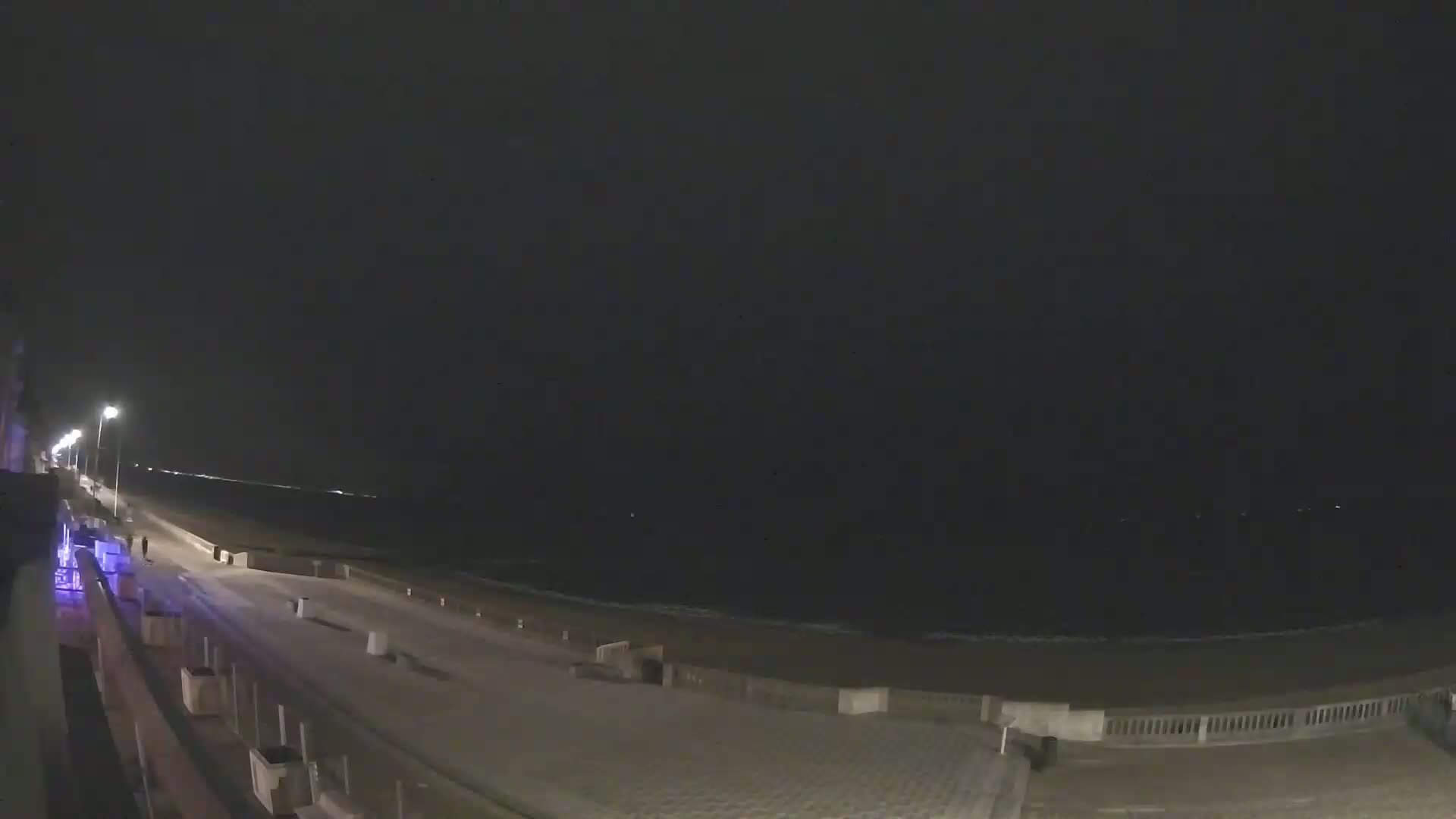 A nighttime view of a snow-covered beachside boardwalk with a calm ocean in the background under a dark sky.