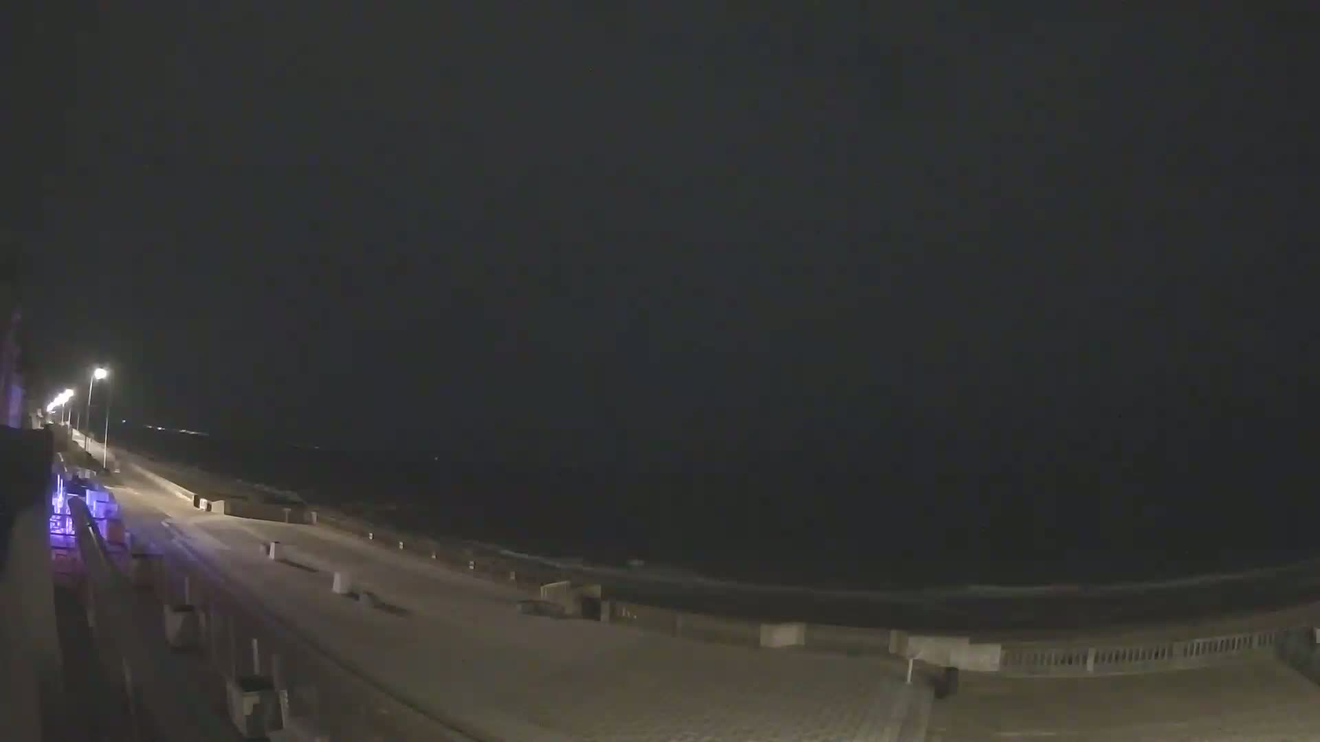 A nighttime view of a dark ocean and a paved beachfront area illuminated by streetlights.
