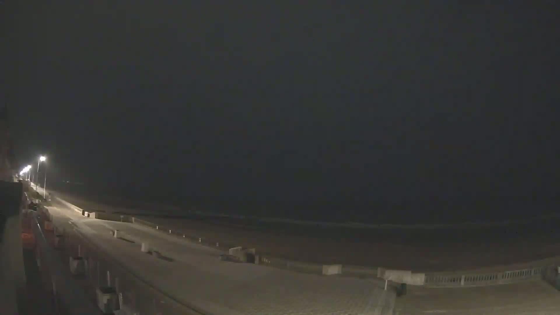 A nighttime view of a mostly empty beach boardwalk with dimly lit streets and a dark ocean in the background.