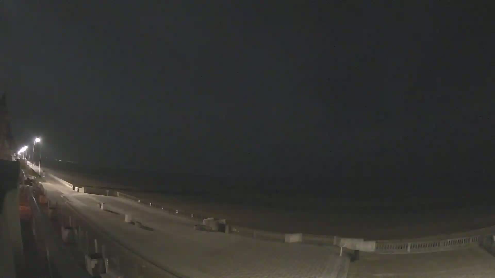 A nighttime view of a dimly lit boardwalk along a beach under a dark, overcast sky.