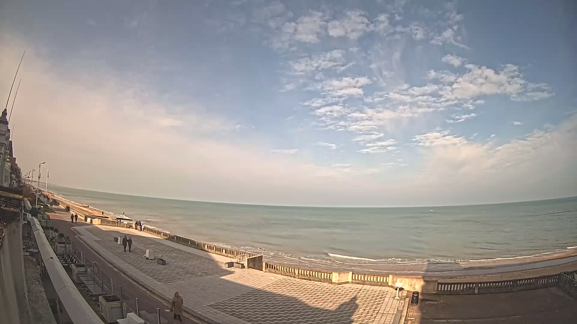 A mostly sunny day reveals a wide expanse of calm ocean meeting a paved beachfront promenade with a few pedestrians.