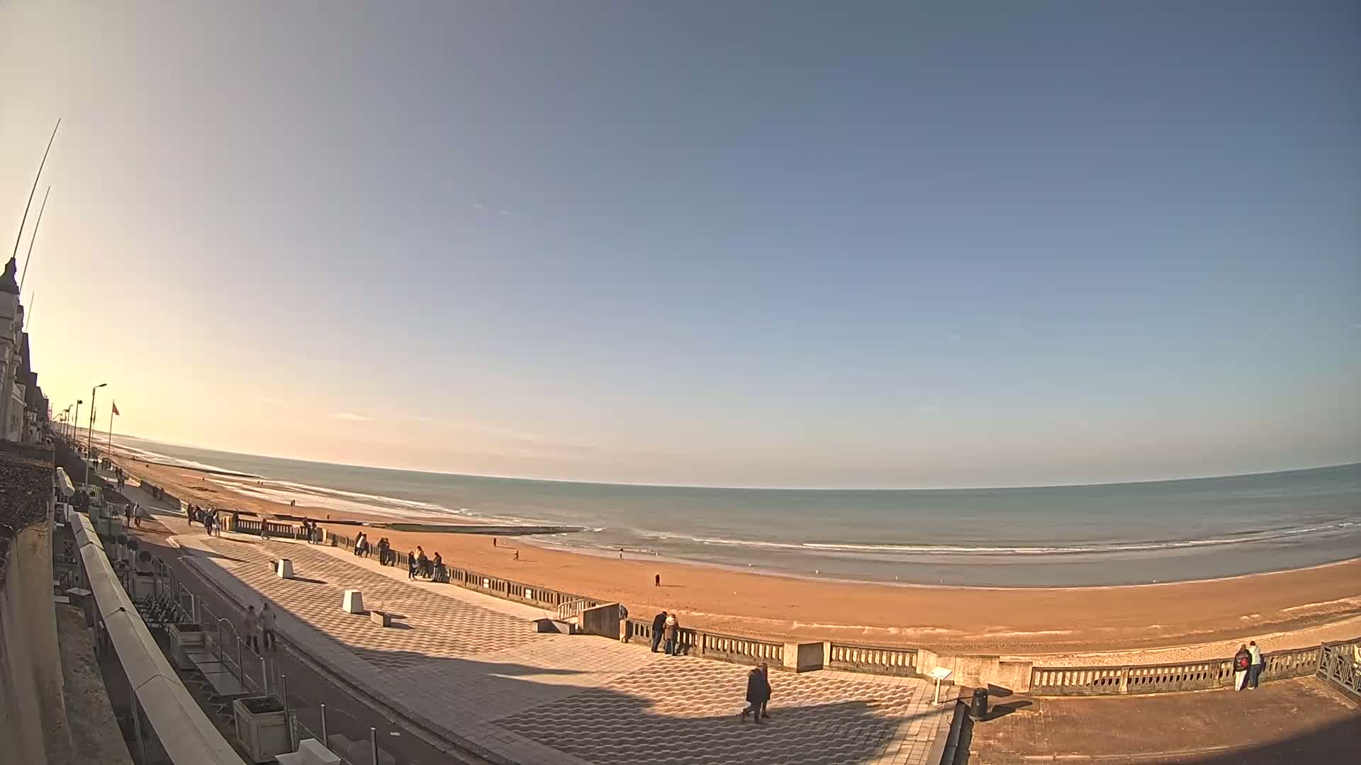 A wide, sandy beach with gentle waves under a clear, bright sky is viewed from an elevated position, showing people strolling along a paved promenade near the ocean.